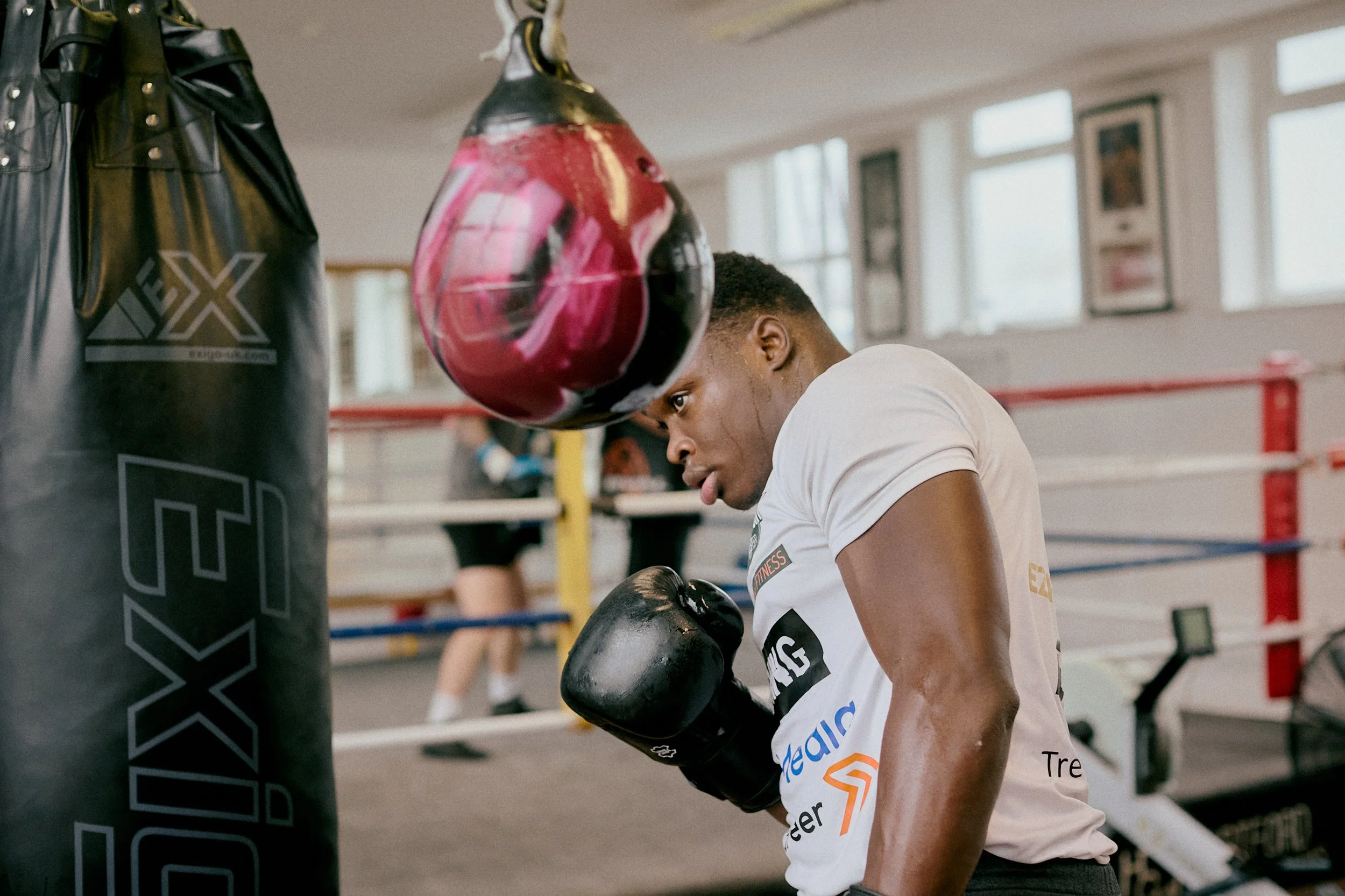 A boxer wearing a white shirt and black gloves training in a gym, leaning against a black punching bag with red and black boxing gloves hanging in front of him.