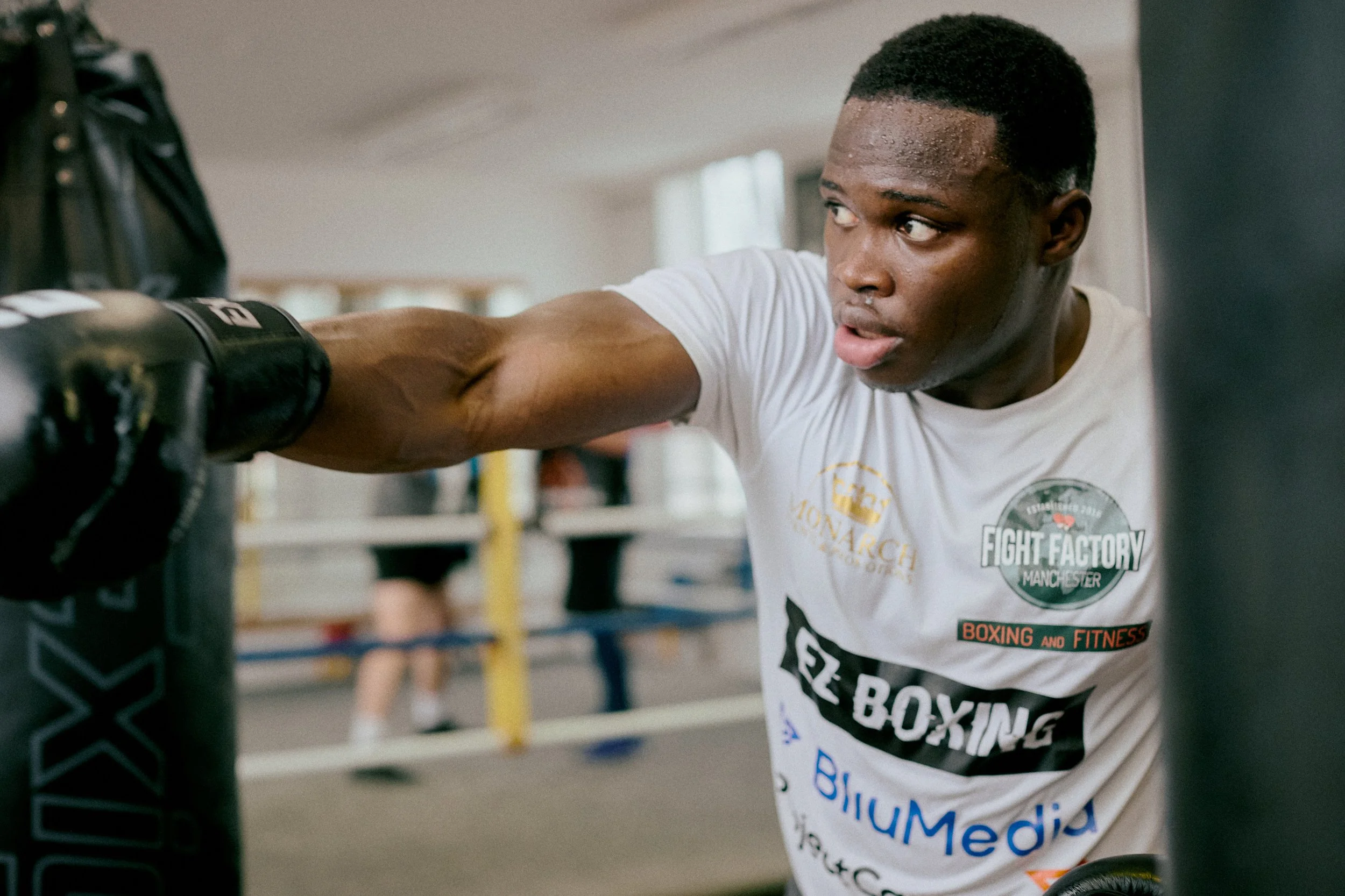 A young man in a white boxing T-shirt is practicing punches on a punching bag at a boxing gym.