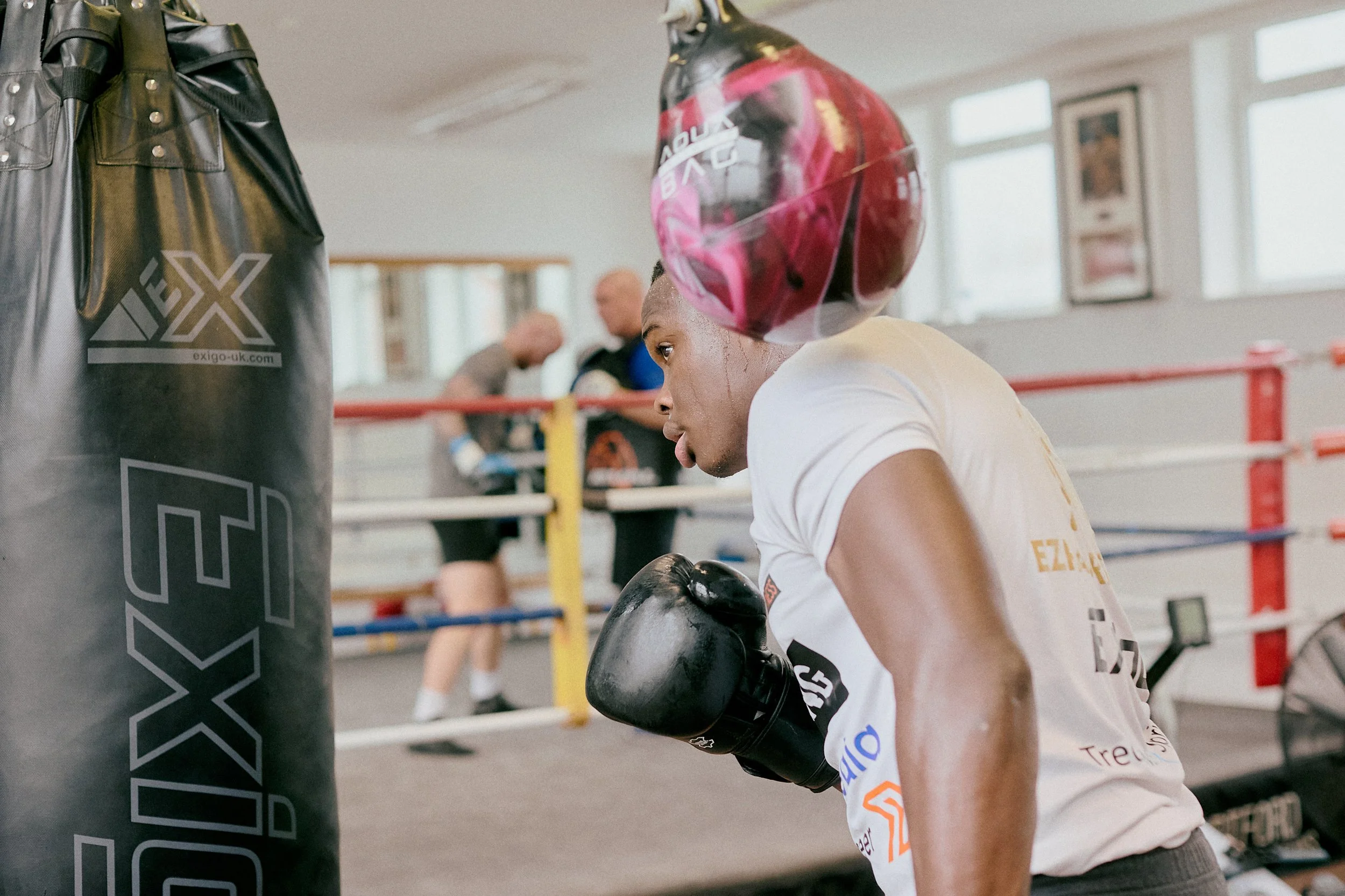 A boxer sparring in a boxing gym, wearing black gloves and a helmet, with a punching bag in the foreground and other people training in the background.
