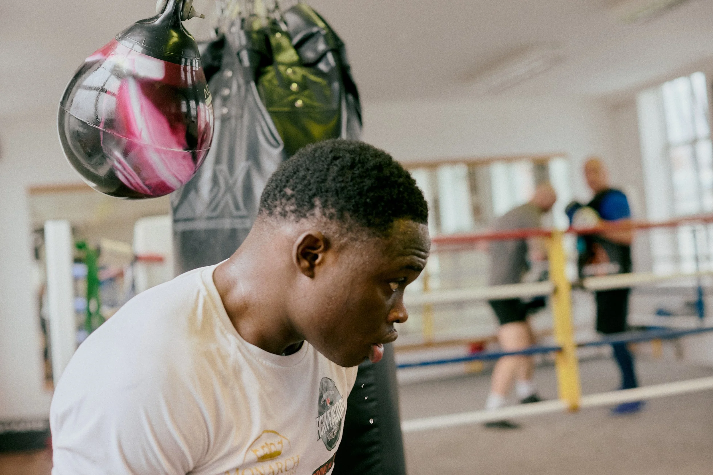 A young man in a white T-shirt resting in a boxing gym, with a boxing ring and two older men in the background, and a punching bag with a colorful punching mitt hanging nearby.