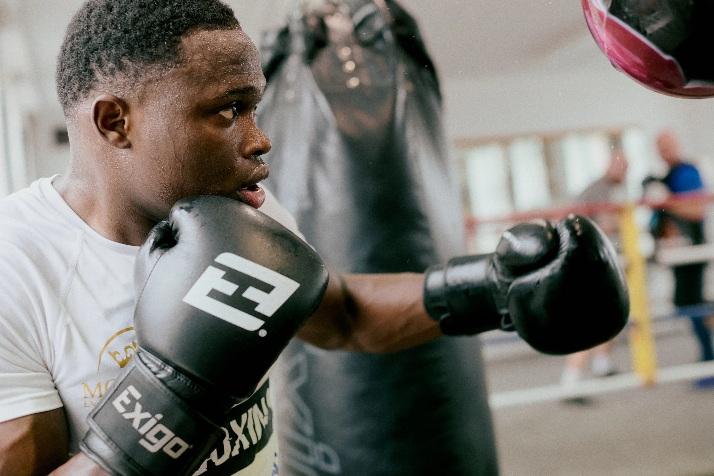A young male boxer in white t-shirt training with a coach in a gym, wearing black boxing gloves, punching a focus mitt held by the coach.