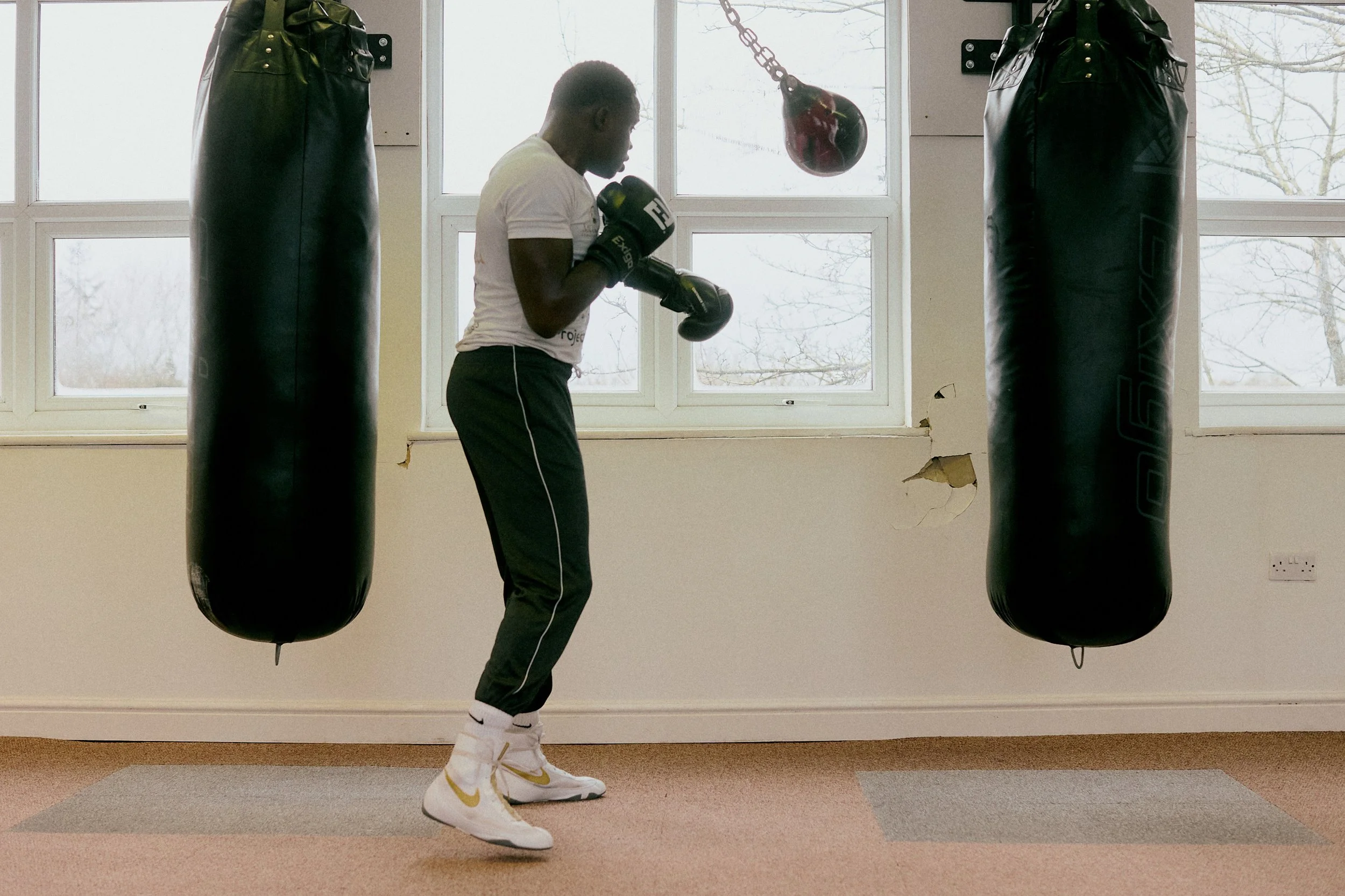 A young man wearing boxing gloves and athletic clothing is practicing boxing in a gym, standing between two hanging punching bags in front of a large window.