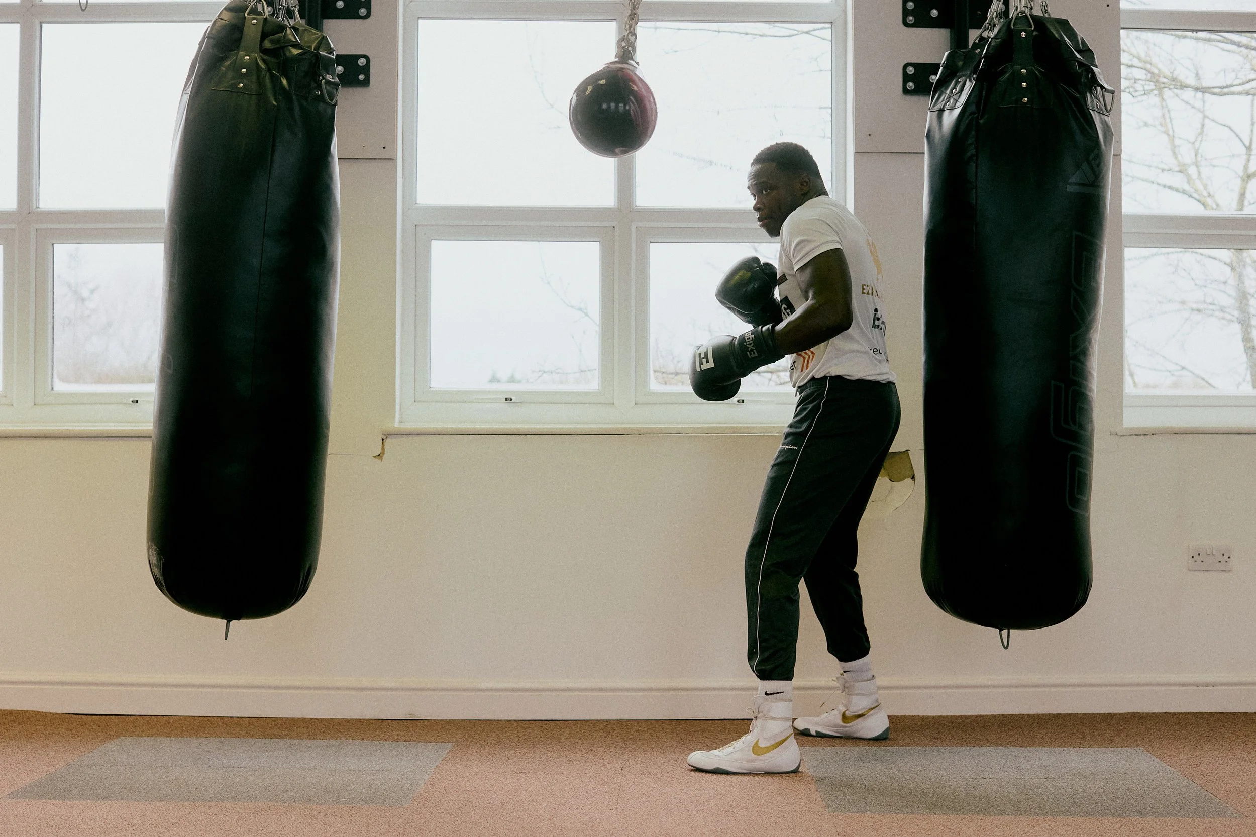 A man training in a boxing gym, wearing boxing gloves, standing in front of two hanging punching bags, with a window behind him