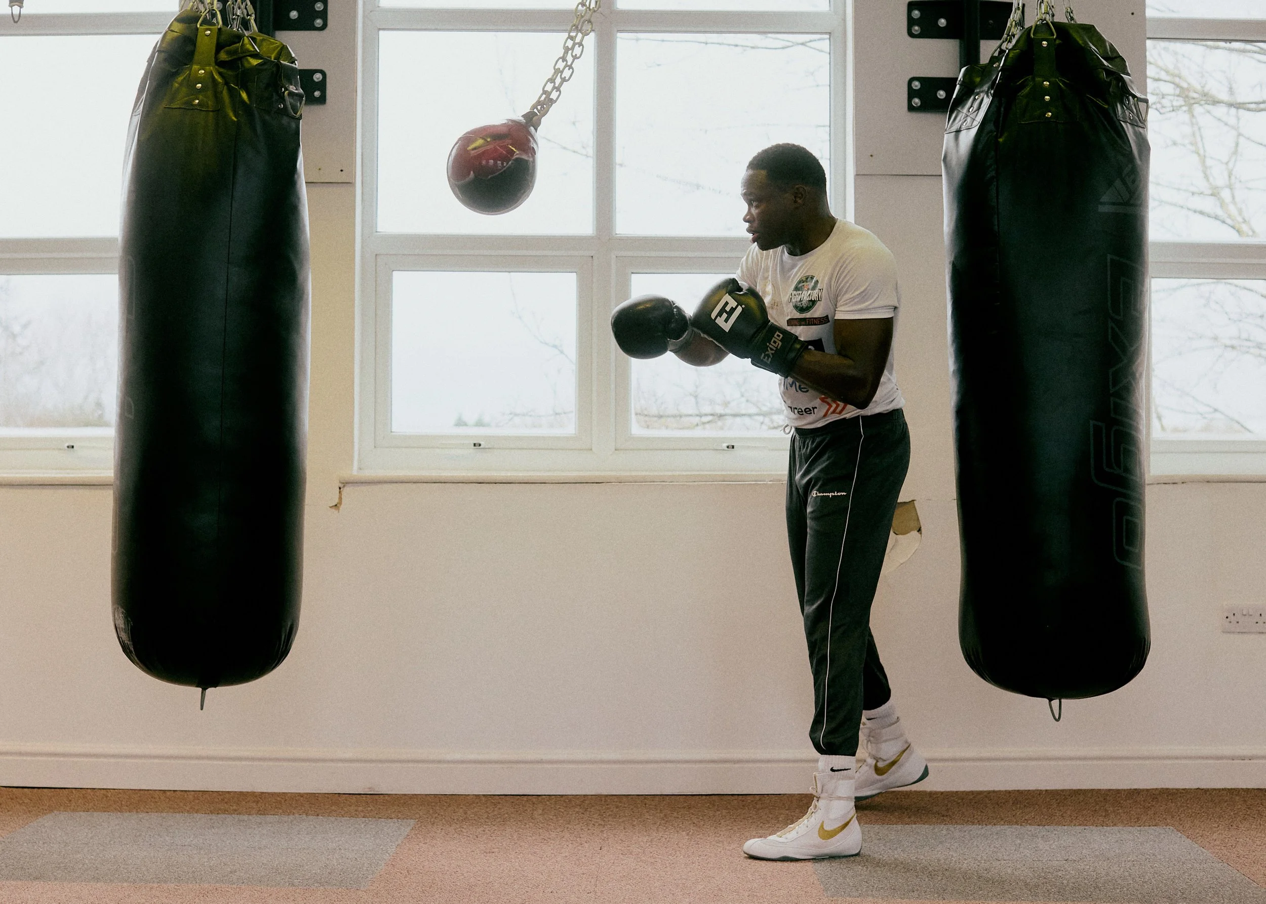 A young man in white t-shirt and black sweatpants with boxing gloves practicing boxing punch in a gym with two black punching bags and a hanging red boxing glove.
