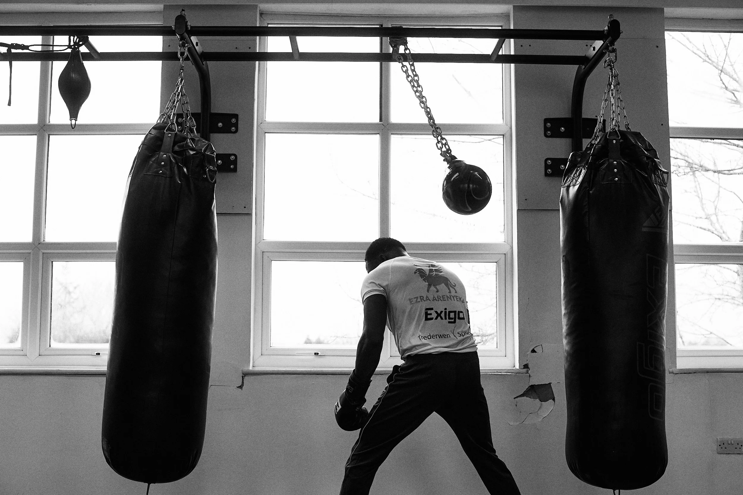 A person practicing boxing in a gym, facing two hanging boxing bags and a punching ball, with large windows in the background.