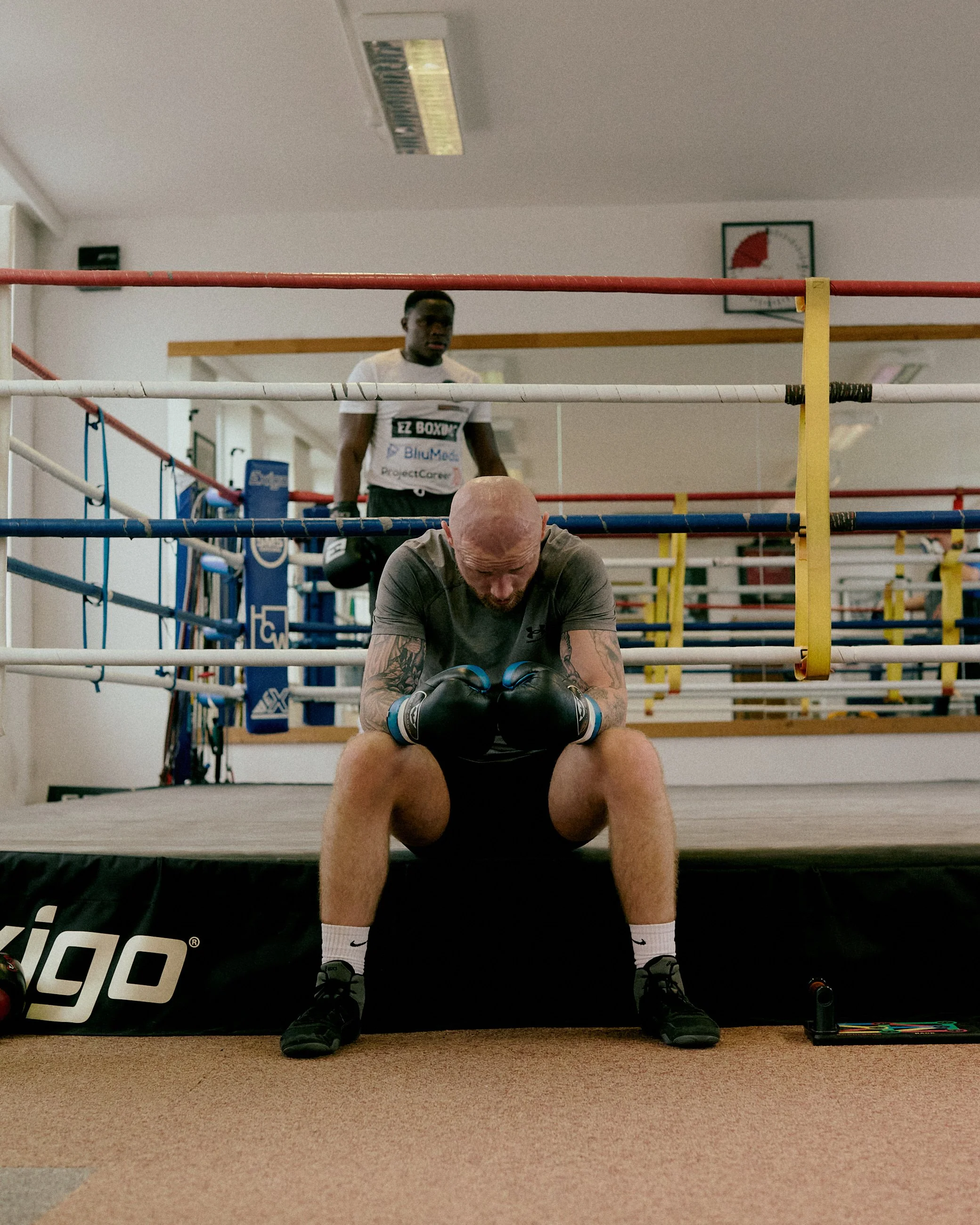 Two boxers in a boxing gym, one sitting on the ring apron with gloves in a praying or resting position, the other standing behind in boxing attire.