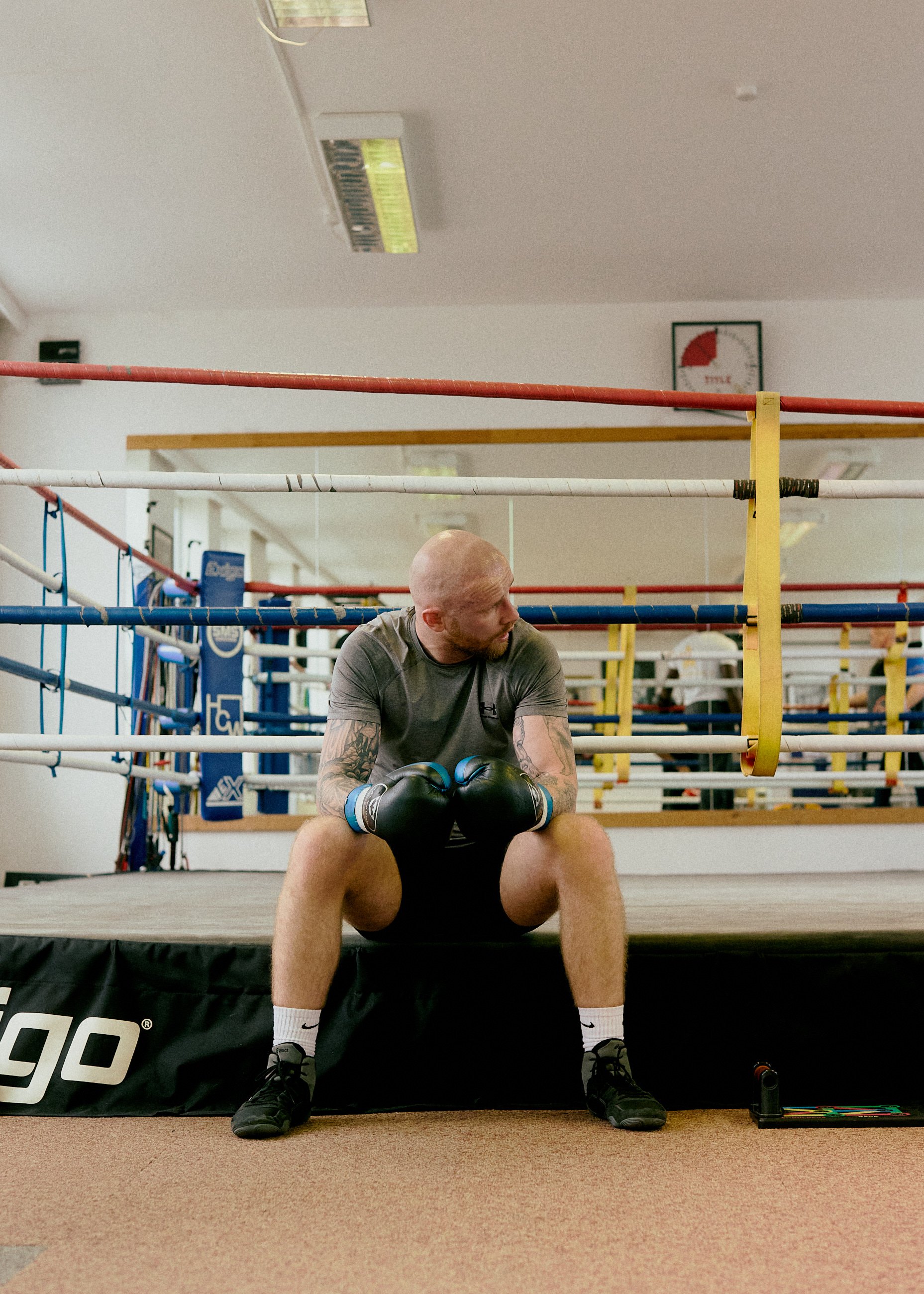 A man with tattoos wearing boxing gloves and athletic clothing sitting on the edge of a boxing ring, looking to the side, inside a gym.