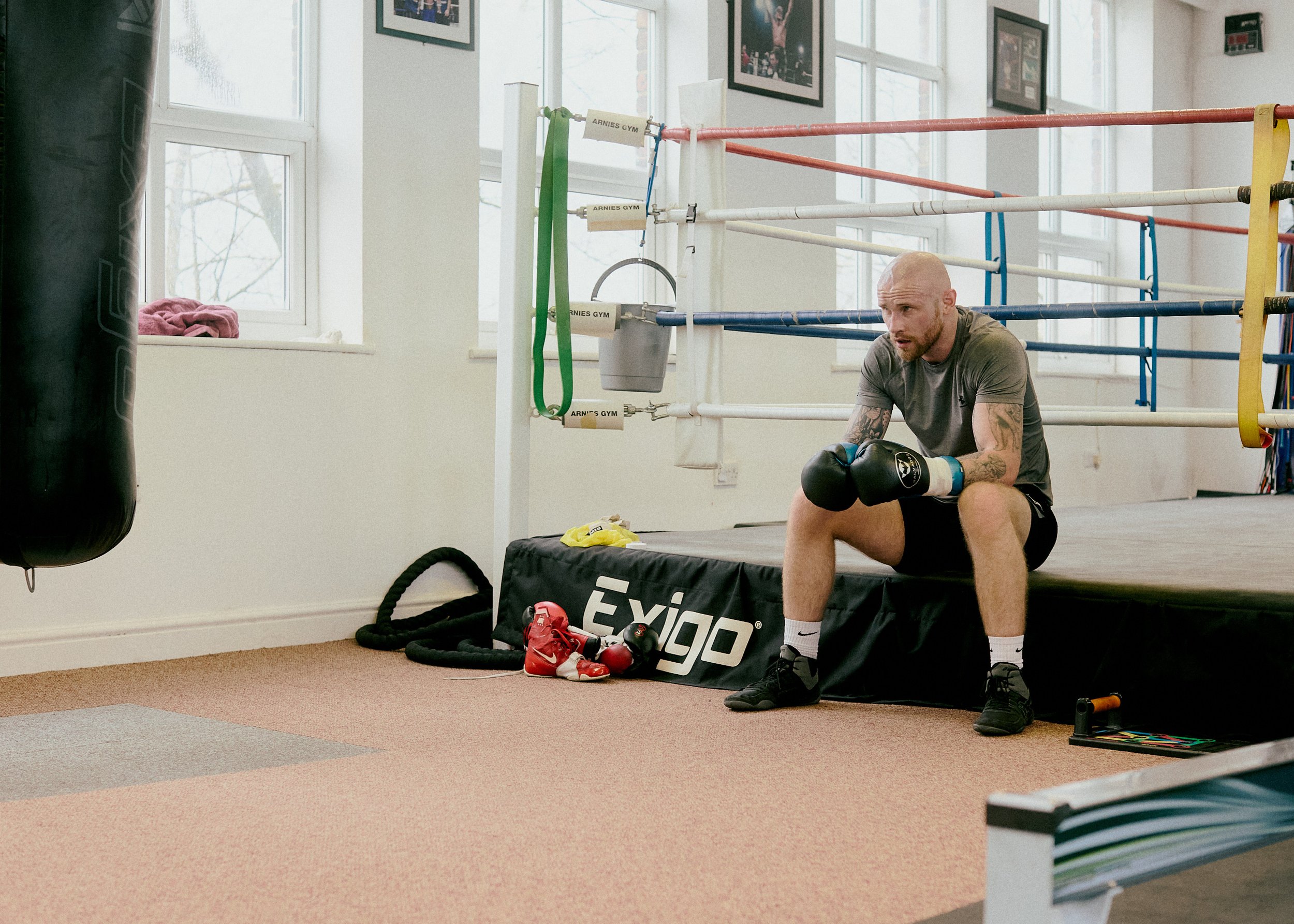A man in boxing gloves sitting on a boxing ring apron in a gym, with boxing gloves and a punching bag nearby.