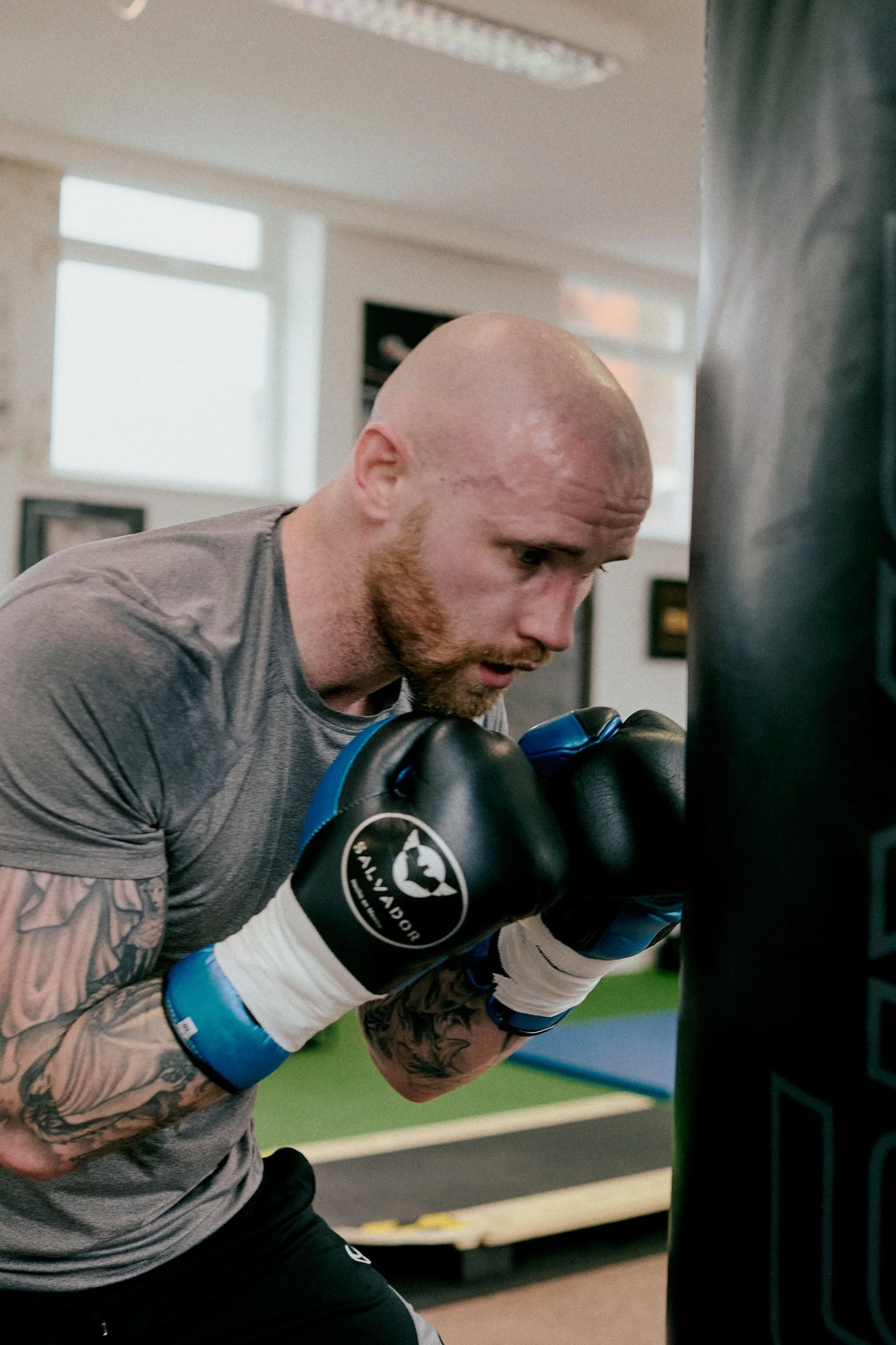 Bald man with tattoos wearing boxing gloves practicing punches on a punching bag in a gym.