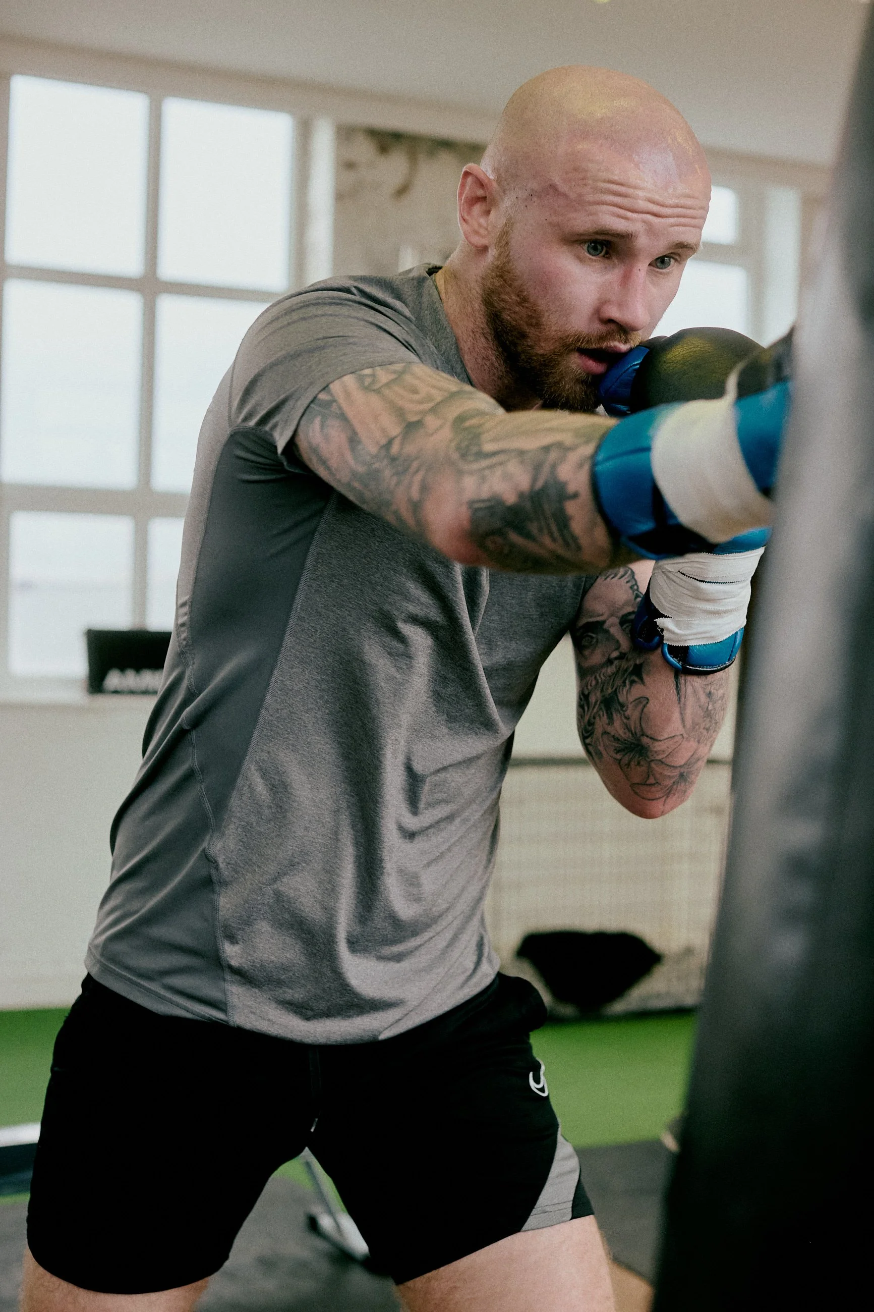 A bald, tattooed man wearing a gray athletic shirt and black shorts is practicing boxing, throwing a punch at a punching bag in a gym.