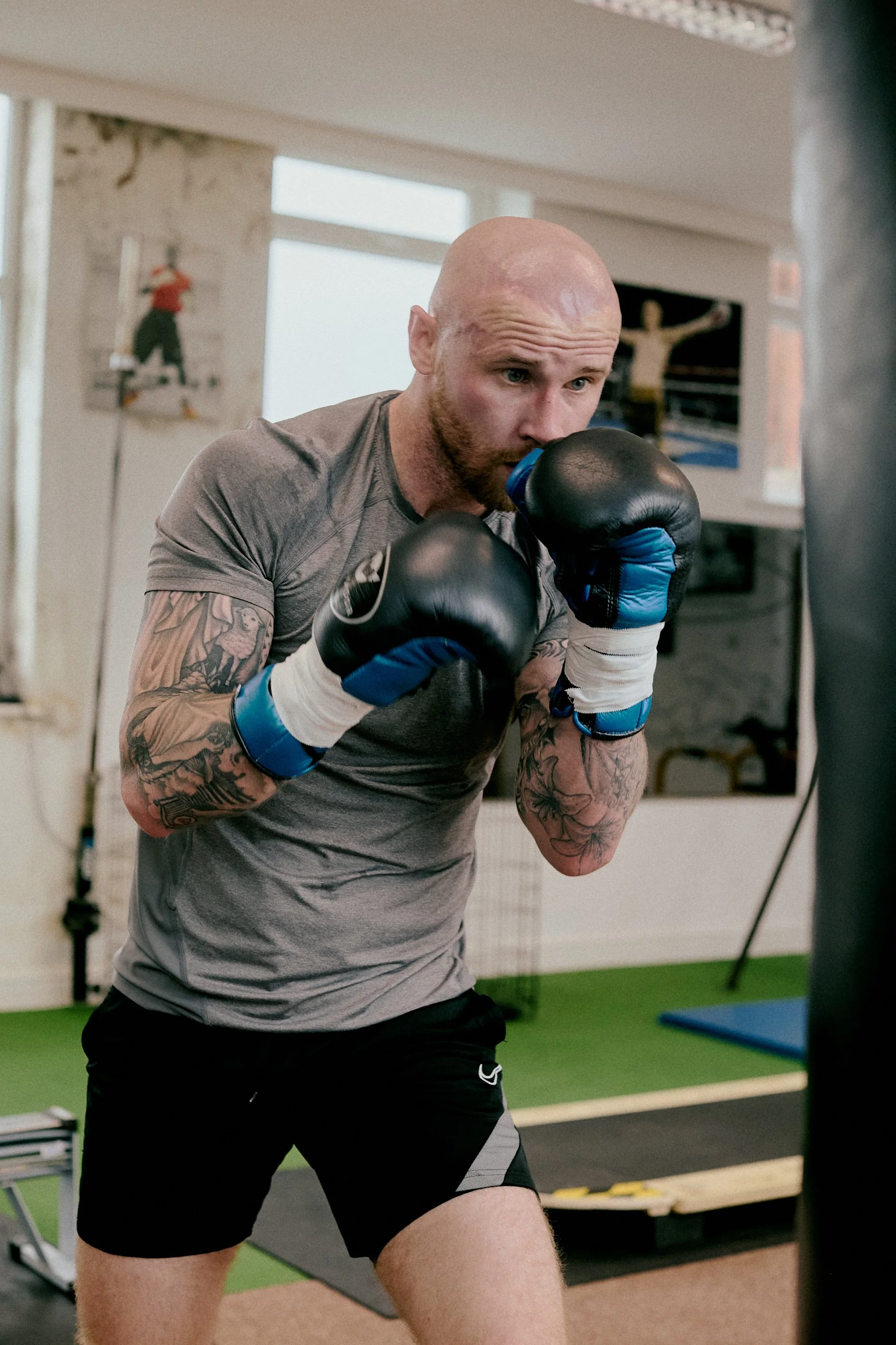 A man with tattoos and a bald head practices boxing with a punching bag indoors.