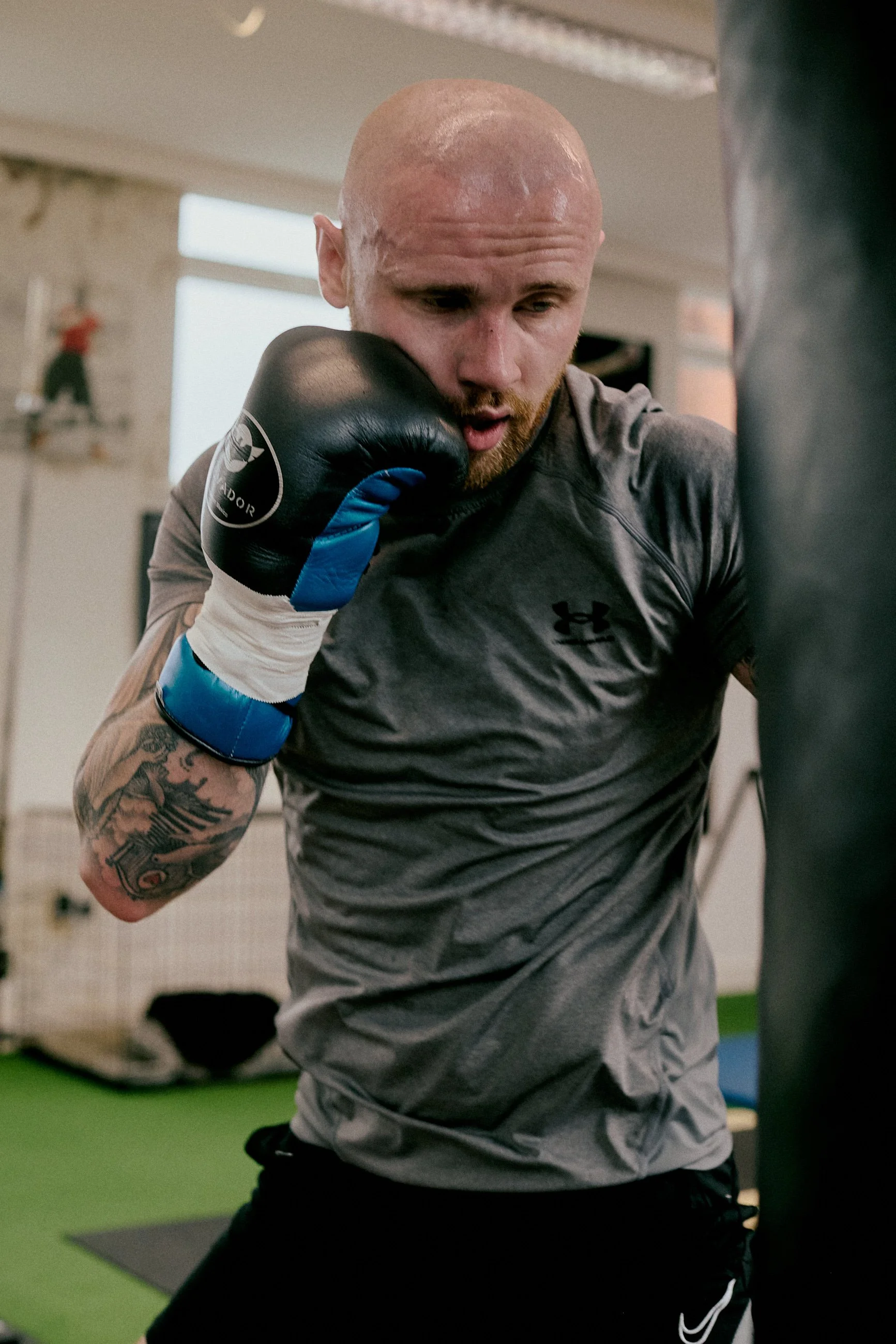 A man in a gray athletic shirt with black Under Armour logo practicing boxing by hitting a black punching bag in a gym. He has a tattoo on his left arm, is bald, and is wearing blue and black boxing gloves.