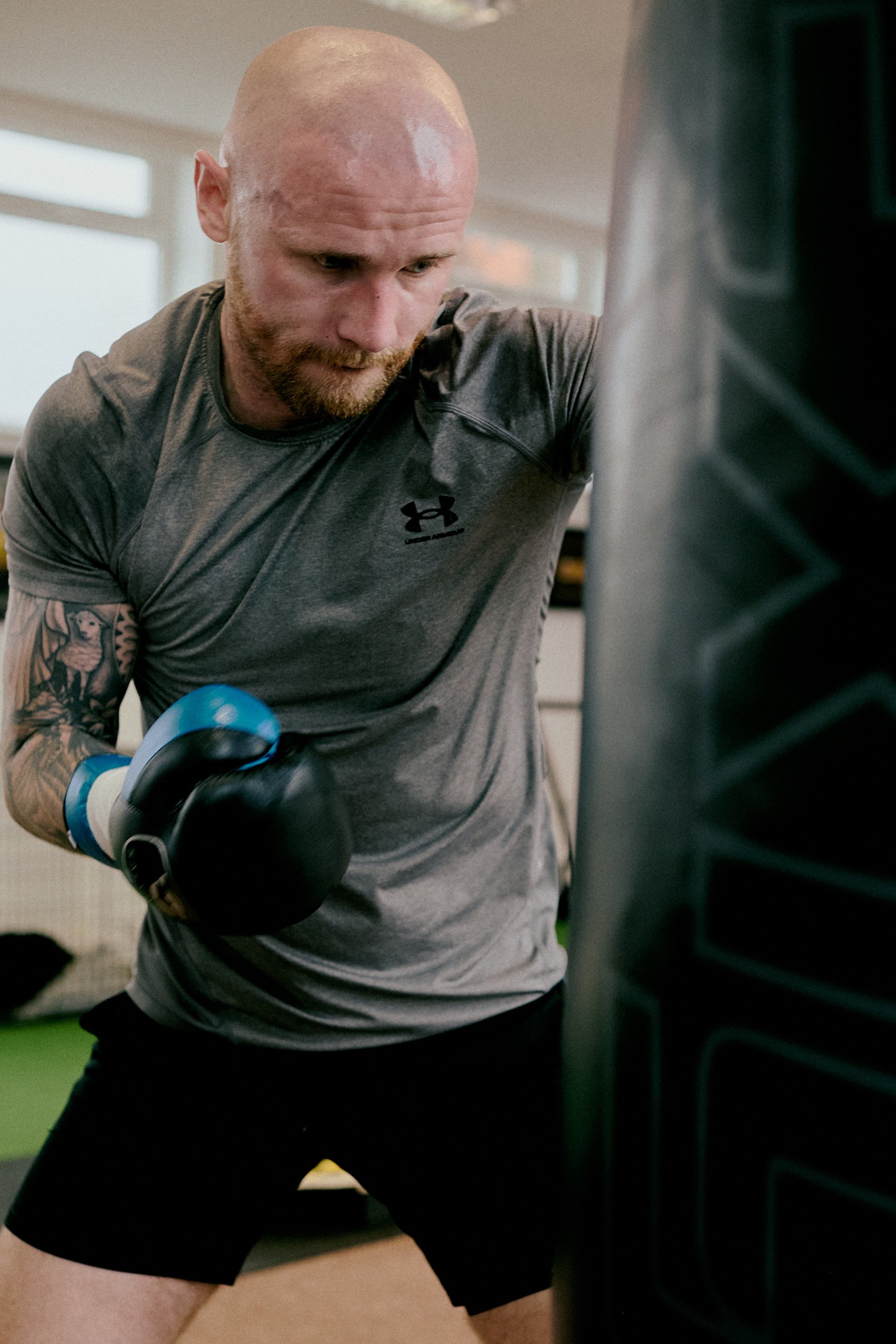 A man with a shaved head and beard wearing a gray athletic shirt and black shorts is boxing a large black punching bag in a gym.