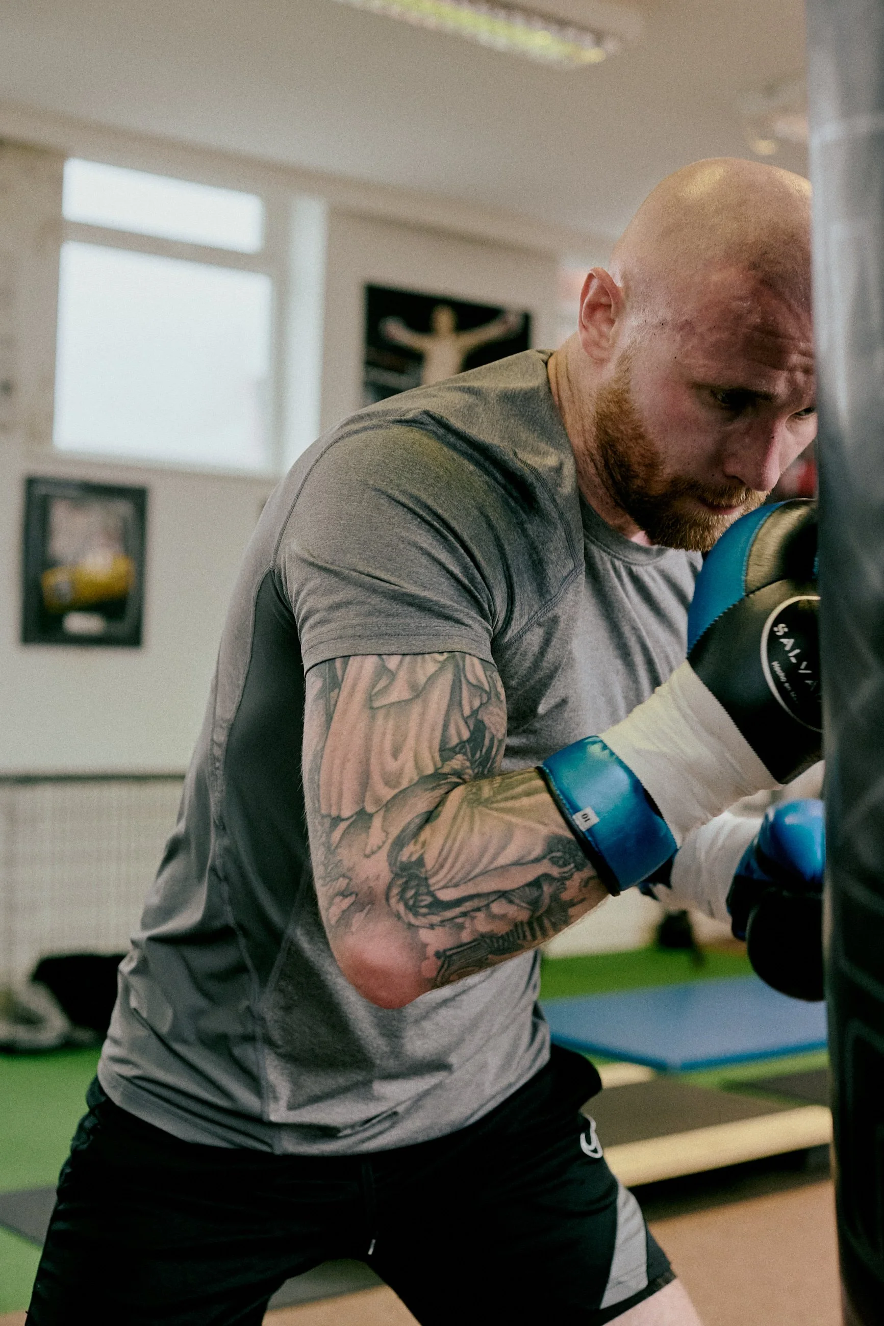 A man with tattoos on his arm, wearing boxing gloves and a gray shirt, boxing at a punching bag in a gym.