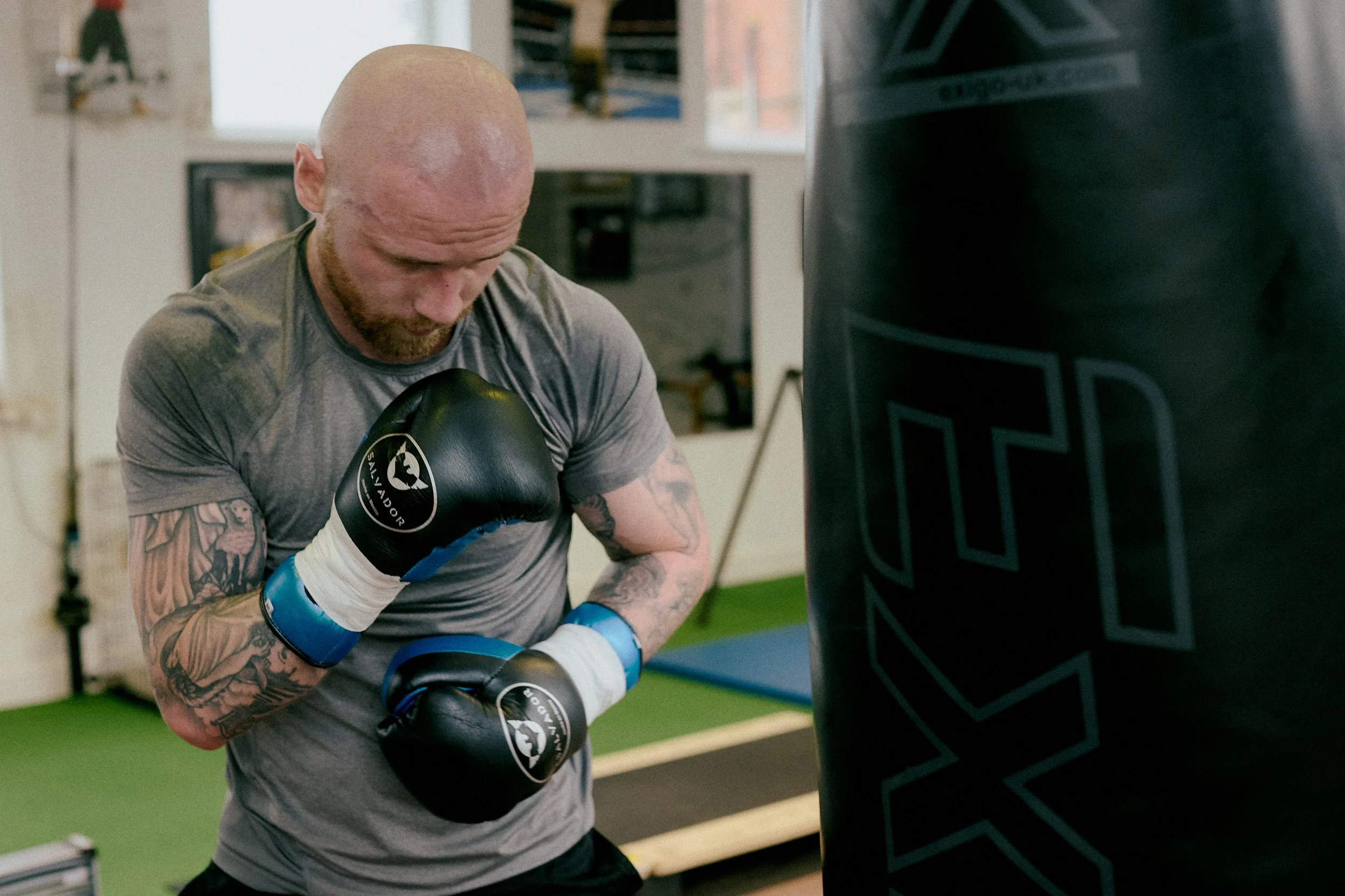 A bald man with tattoos on his arms wearing a gray t-shirt and boxing gloves practicing punch on a punching bag in a gym.