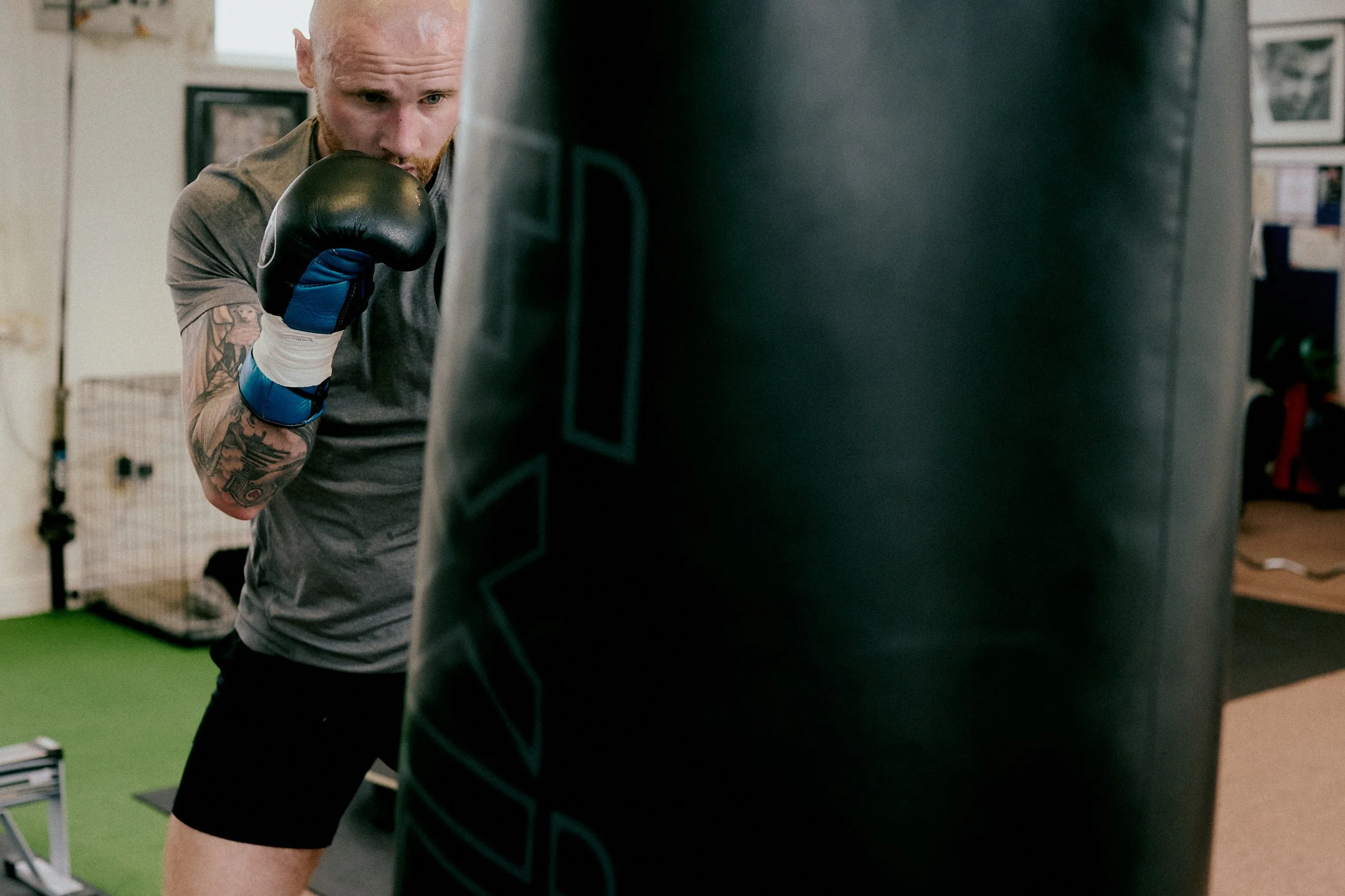 A man with tattoos on his arm wearing a gray shirt and black shorts is training with a punching bag, wearing black boxing gloves with blue accents. The man is in a boxing stance, looking focused on the bag in a gym setting.