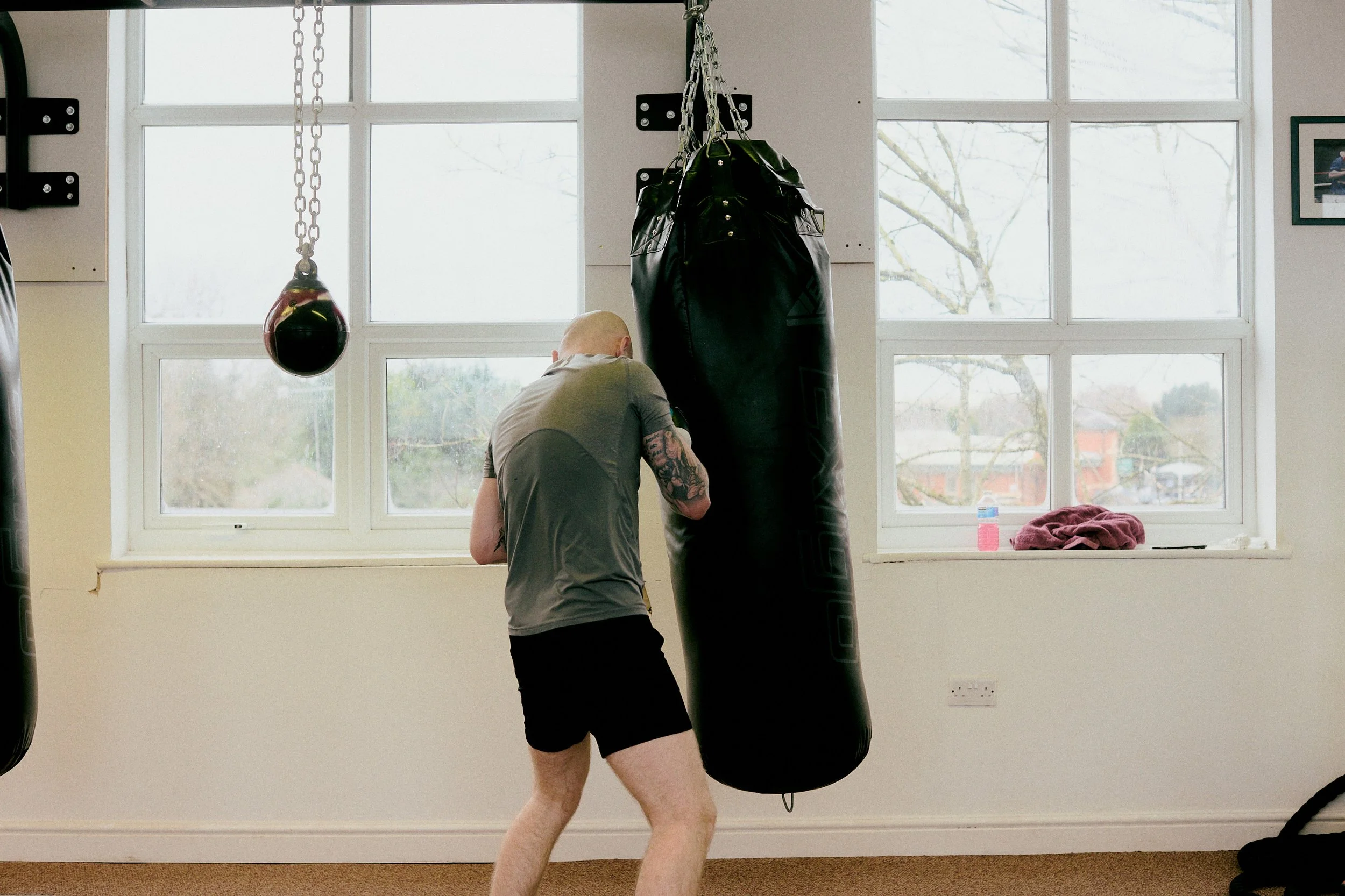 A man in workout clothes punches a heavy boxing bag in a gym with large windows.