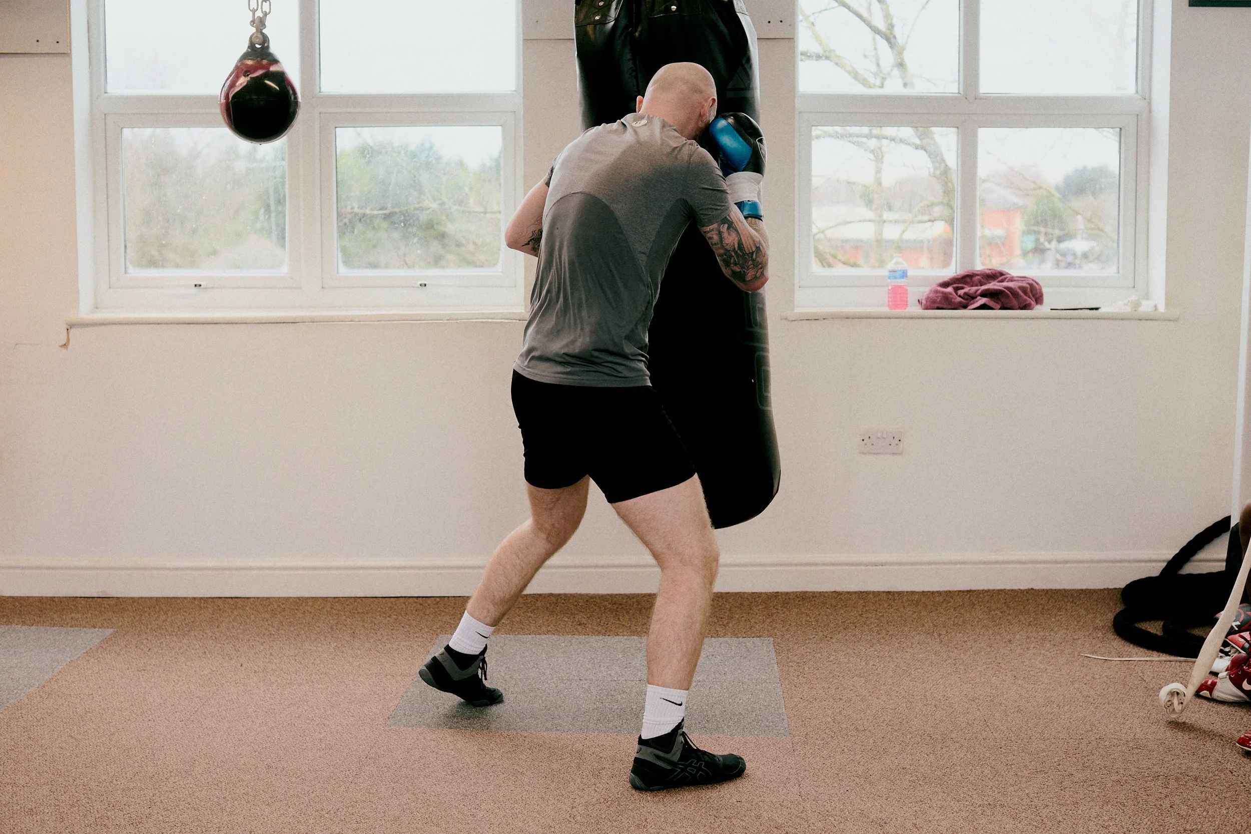 Man with a bald head, wearing a gray shirt, black shorts, and boxing shoes, is practicing boxing on a hanging punching bag in a room with beige carpeting and white framed windows.