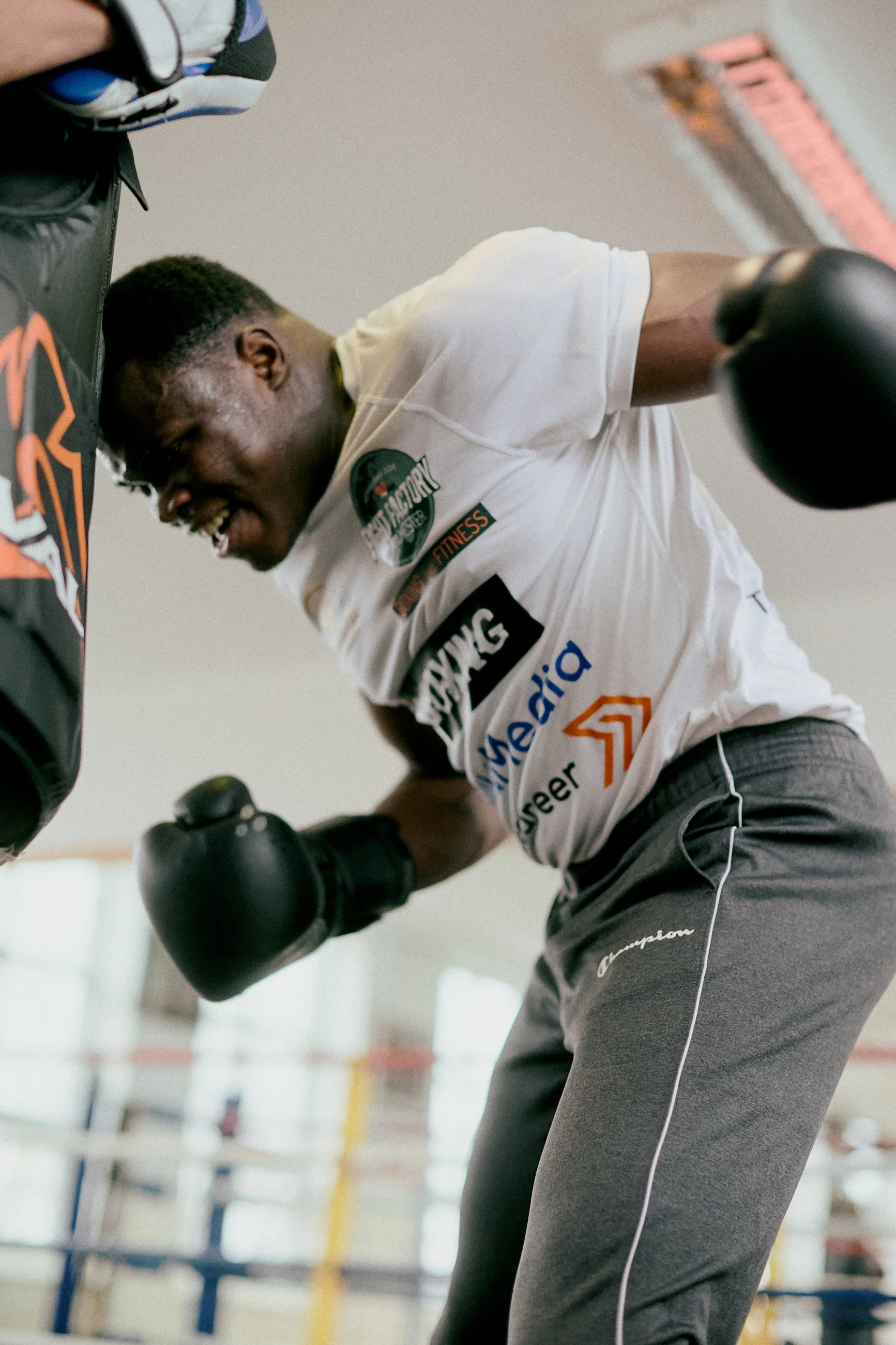 A young man in a fitness gym wearing boxing gloves, a white t-shirt with multiple logos, and dark sweatpants, smiling as he trains.