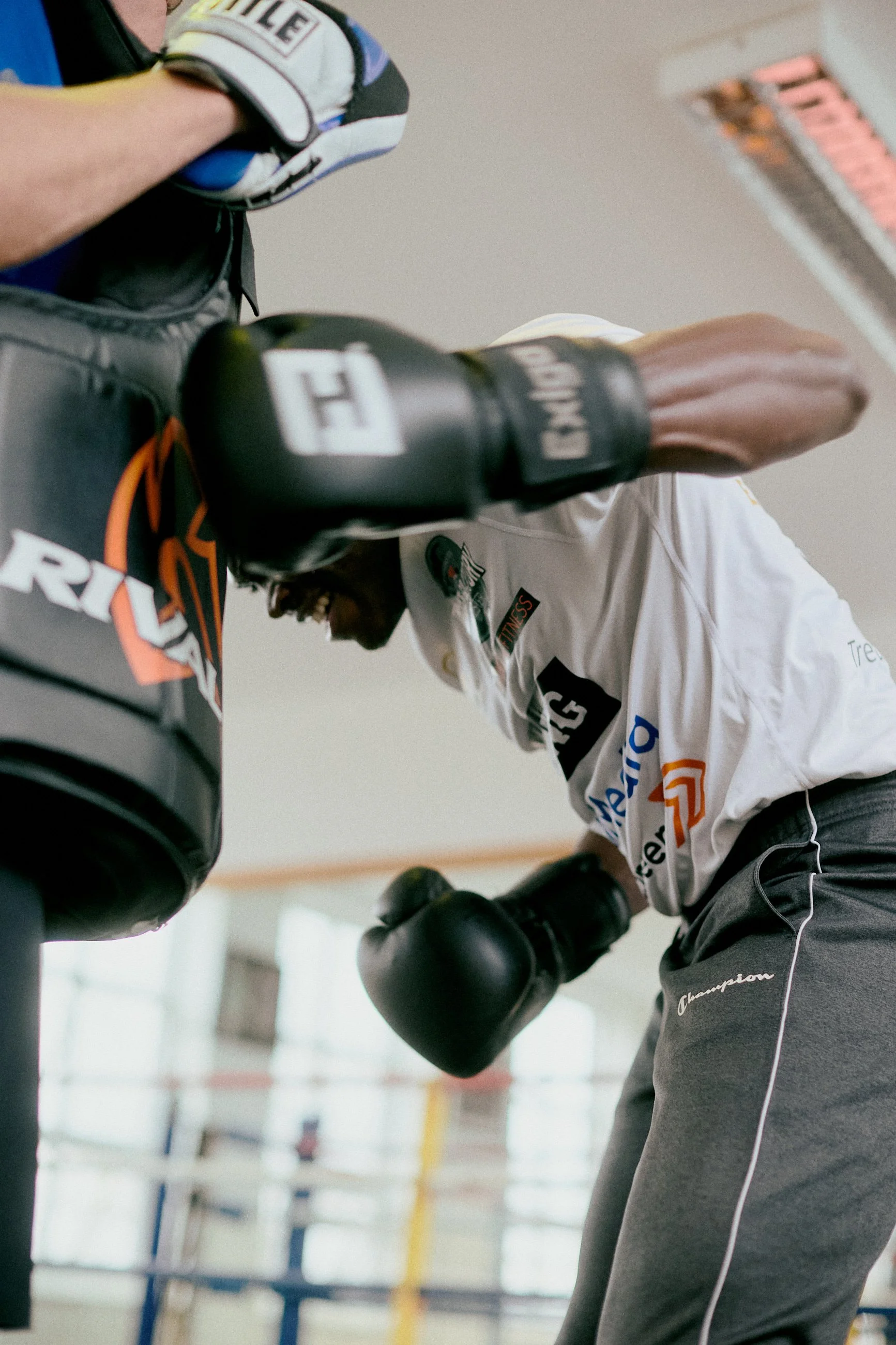 A male boxer in gray sweatpants and a white training shirt practicing punches with black boxing gloves in a gym.