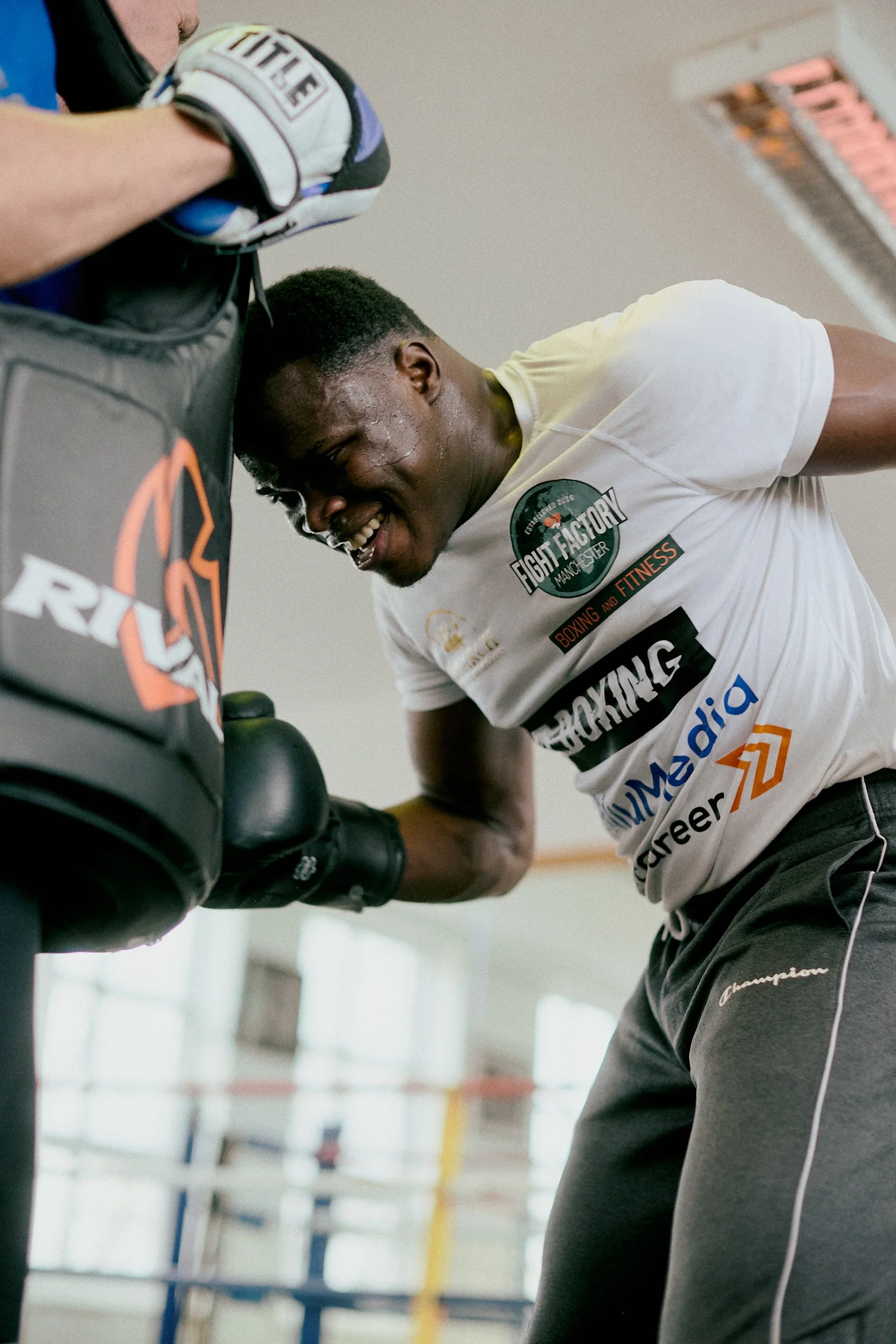 A young African American man is boxing in a gym, smiling and leaning against a boxing bag, wearing a white t-shirt with various logos and black athletic pants.