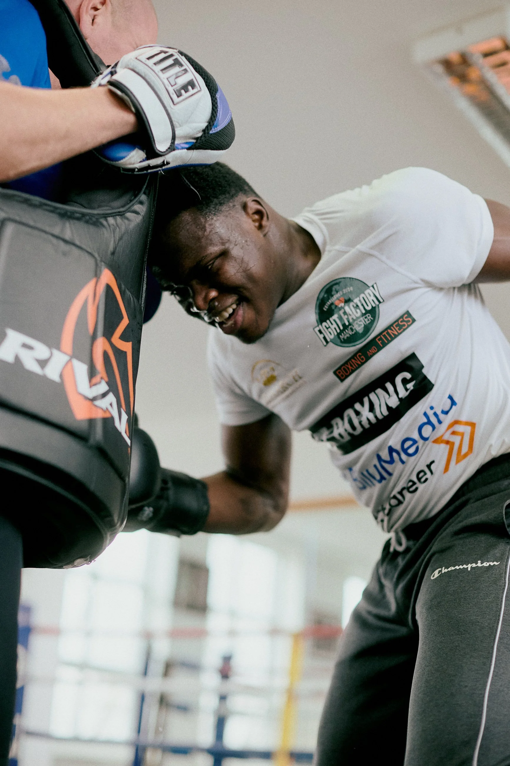 Young male boxer in a gym with a white t-shirt and black shorts leaning on a punching bag, smiling and sweating.