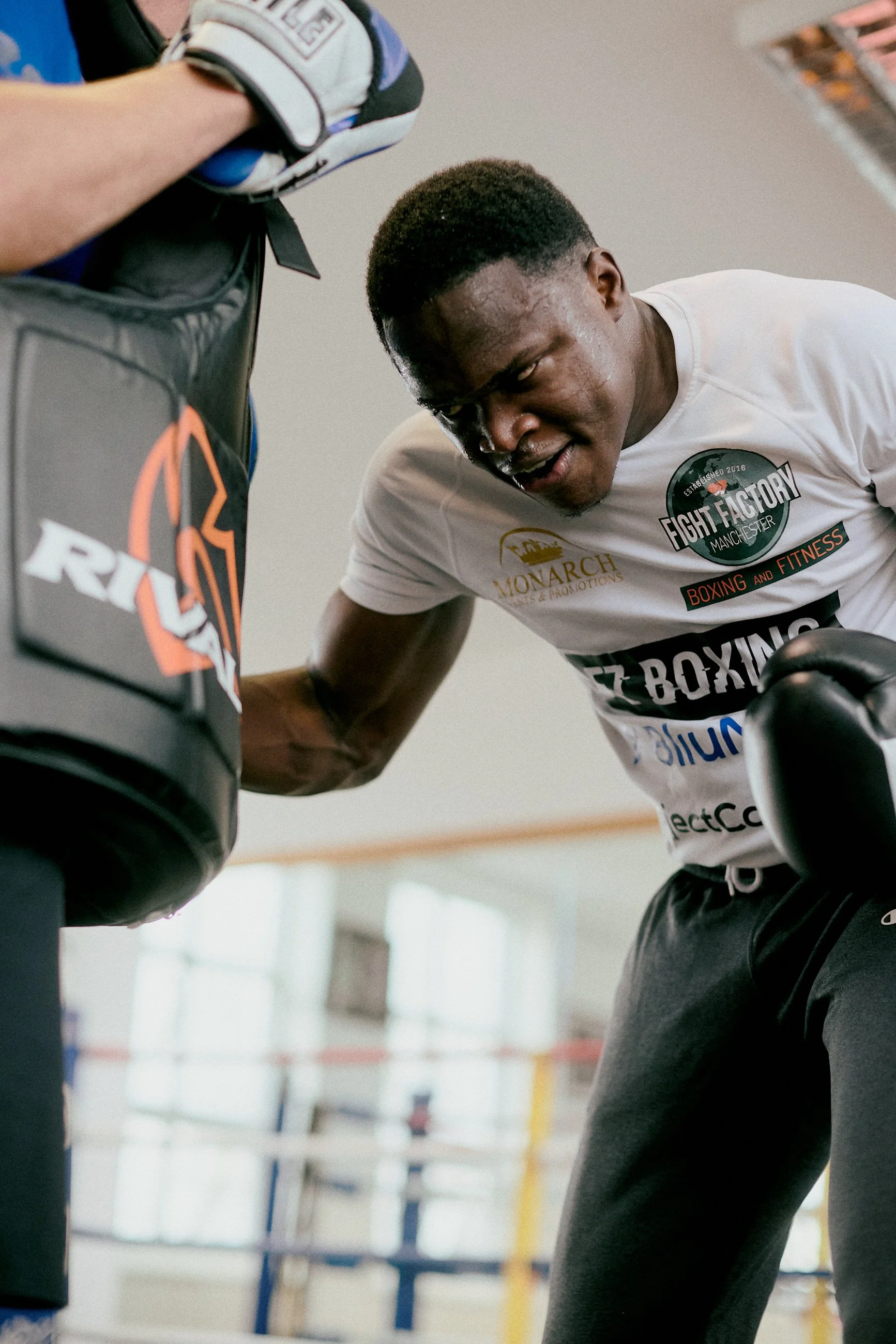 A young man training with a boxing punching bag in a gym, wearing a white t-shirt with various logos, including 'Fight Factory' and 'Boxing and Fitness.' He appears focused and is leaning close to the bag, possibly punching or working out.