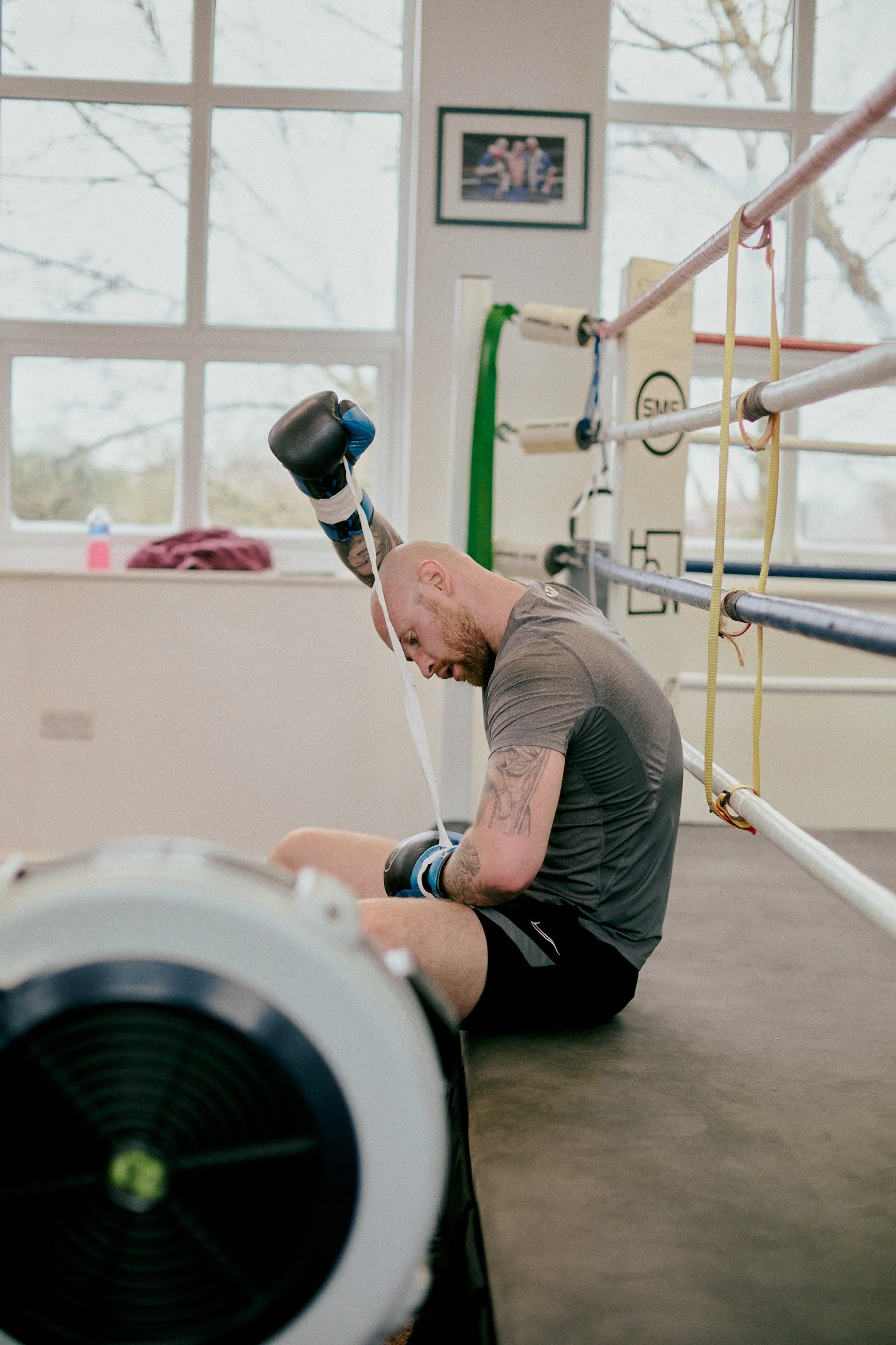 A male boxer sitting on the floor of a boxing gym, resting after a workout, with boxing gloves and gym equipment visible around him.
