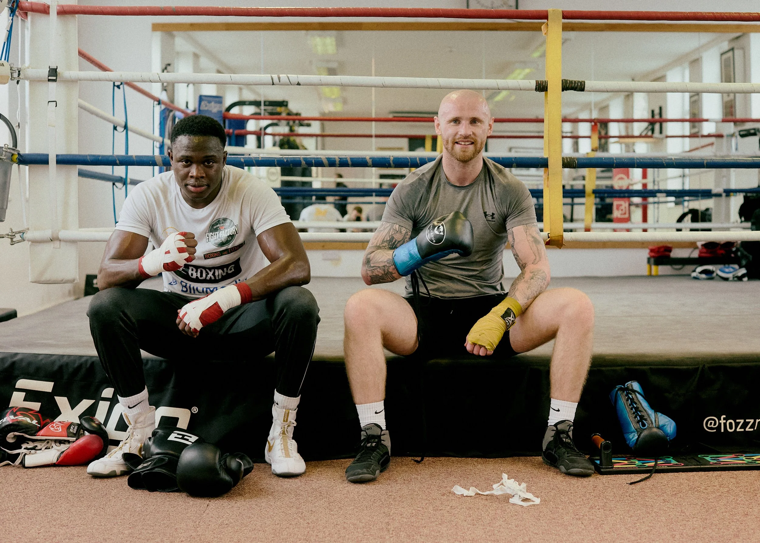 Two male athletes sitting on a boxing ring platform, wearing boxing gloves and athletic gear, in a gym with boxing rings and equipment in the background.