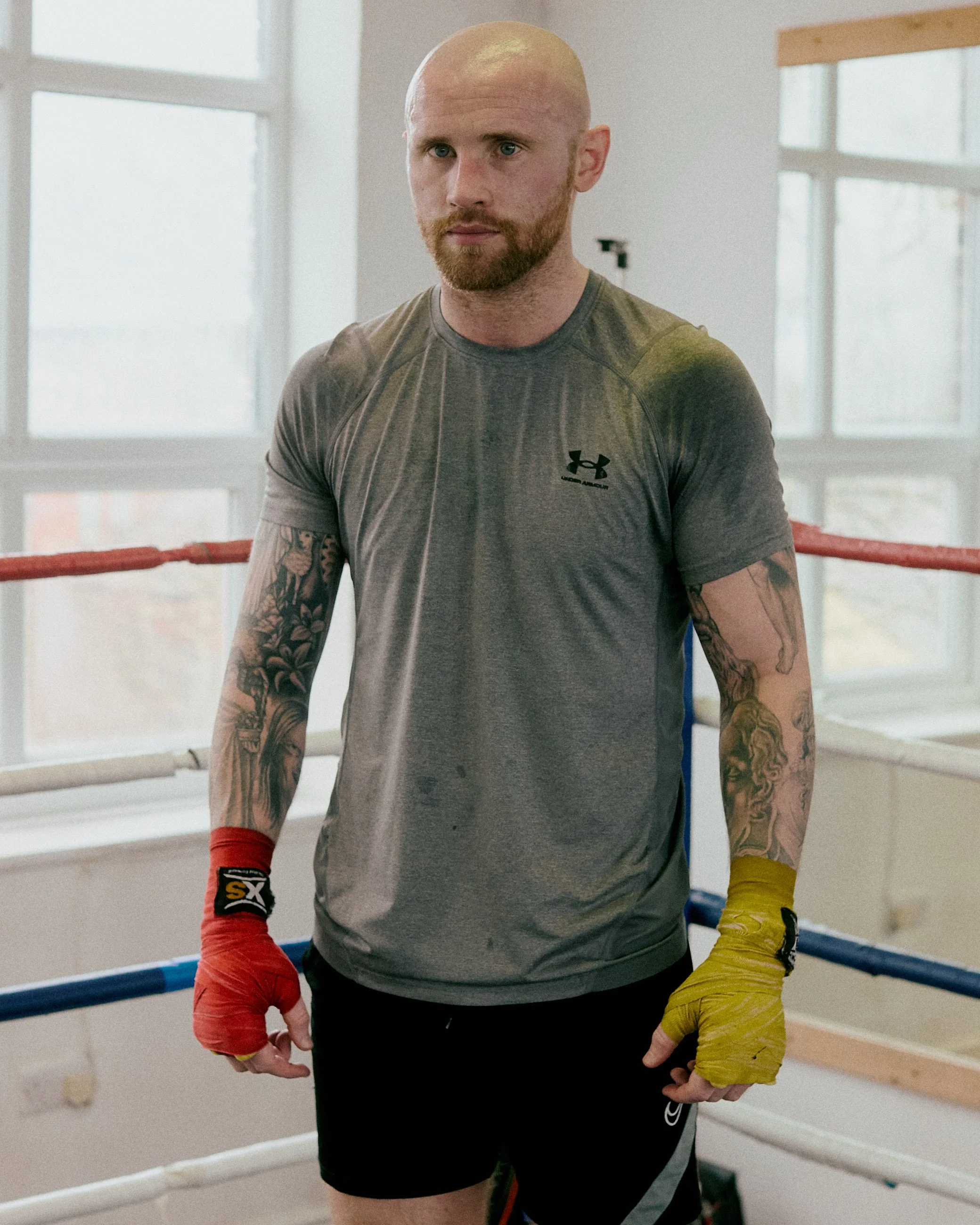 A male boxer with tattoos on his arms, wearing a gray athletic shirt with the Under Armour logo, standing inside a boxing ring with red and blue corner pads, and yellow hand wraps on his right hand.