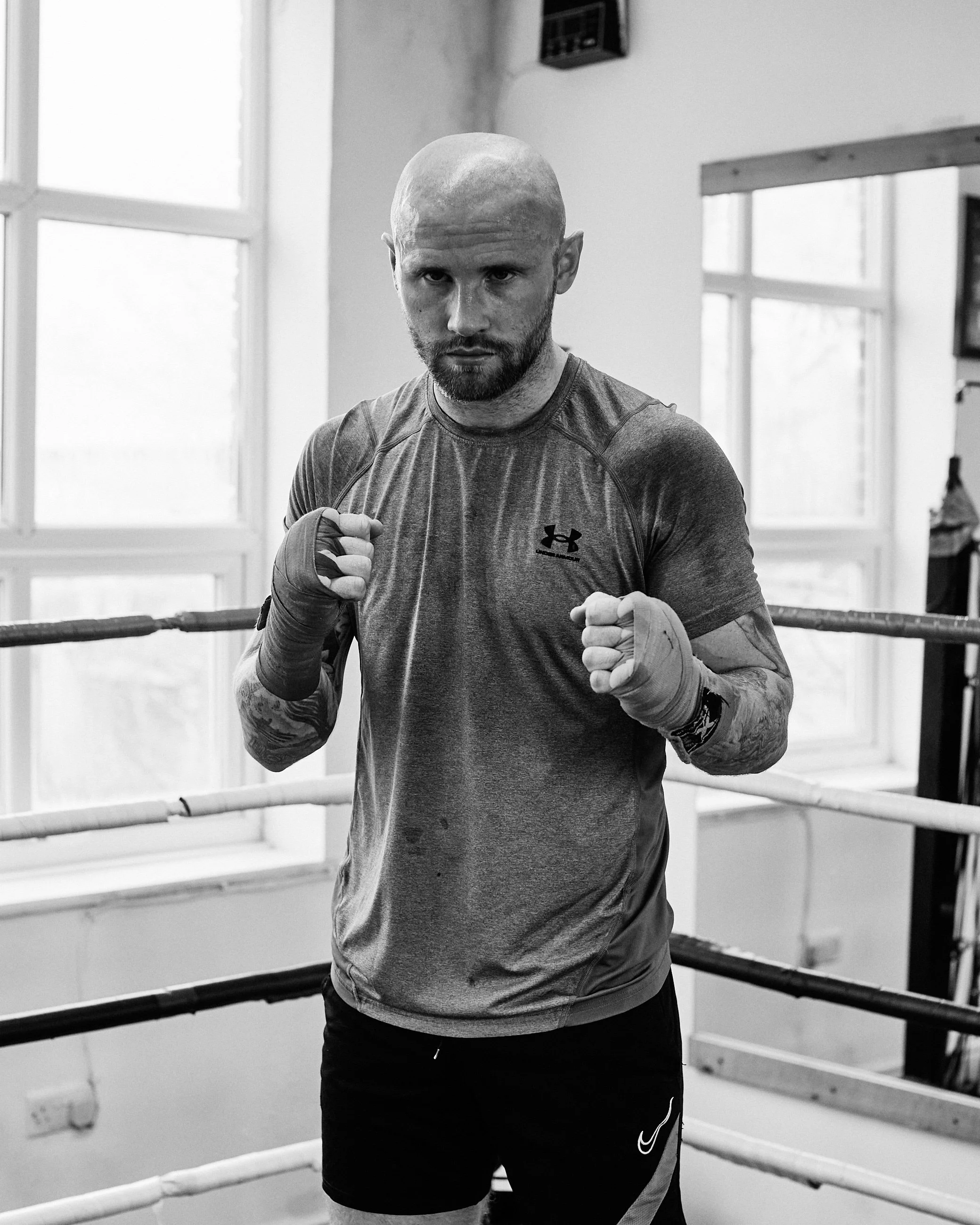 A male boxer posing in a boxing ring with fists up, wearing athletic gear, in a gym with large windows in the background.