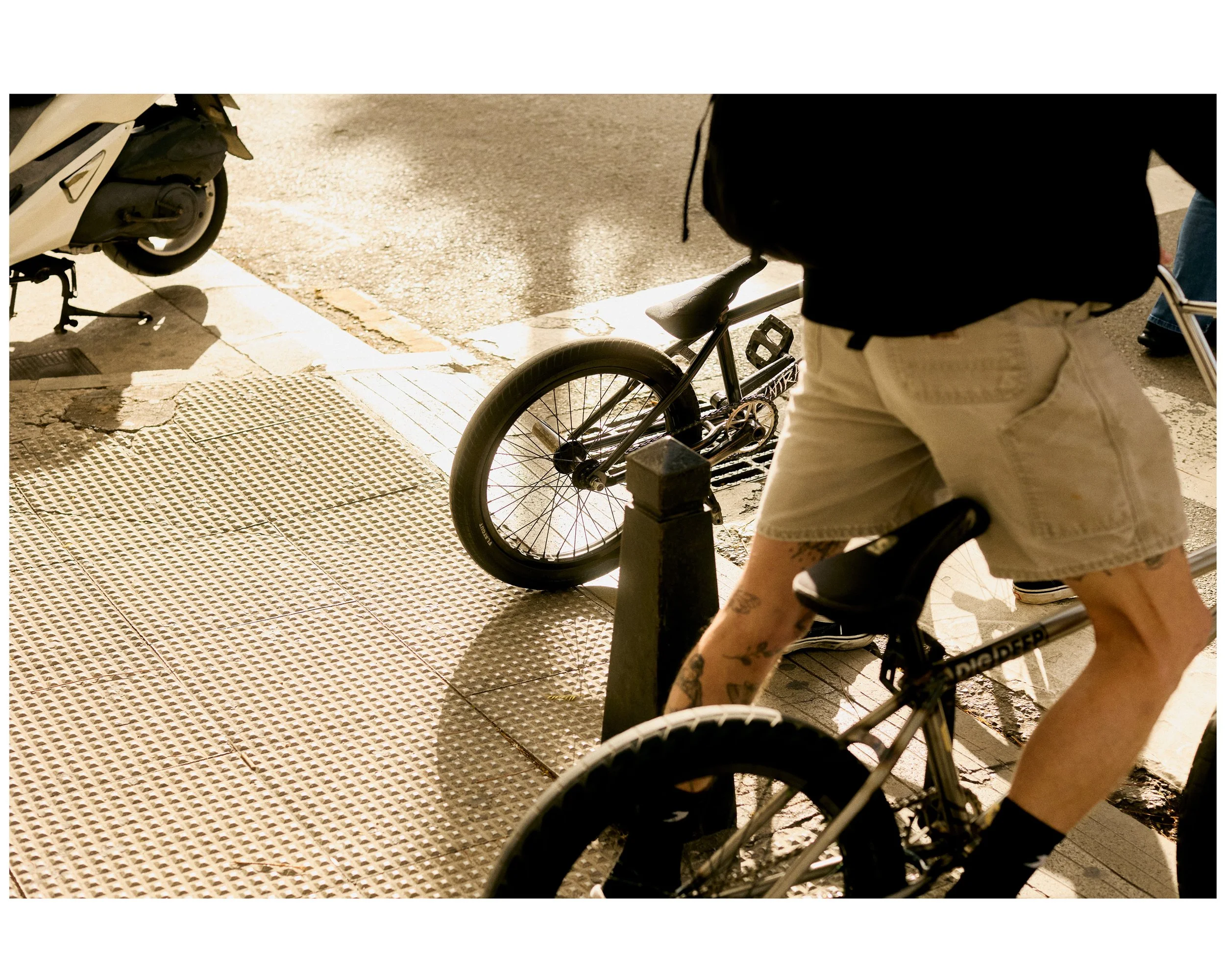 Person wearing shorts riding a bicycle on city sidewalk, with a parked bicycle and scooter nearby, and sunlight casting shadows.