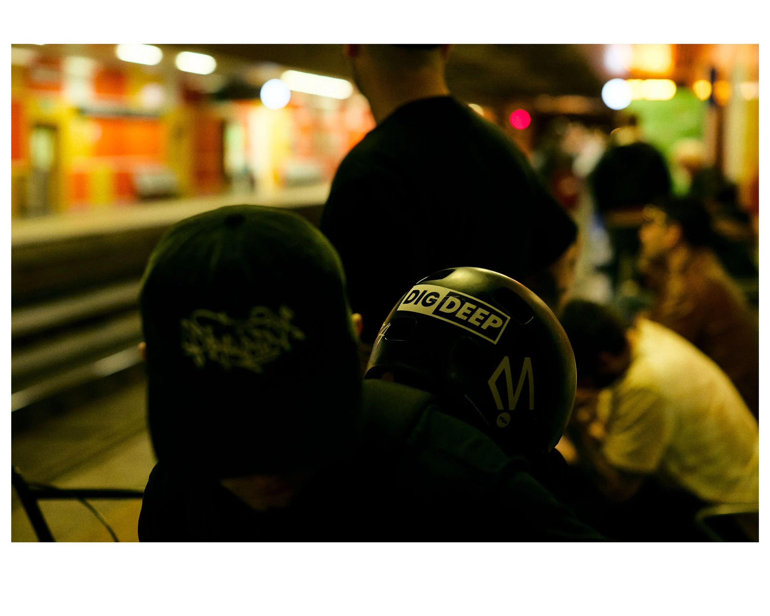People sitting outdoors at night, some wearing hats and helmets, socializing and waiting in line at a brightly lit area.