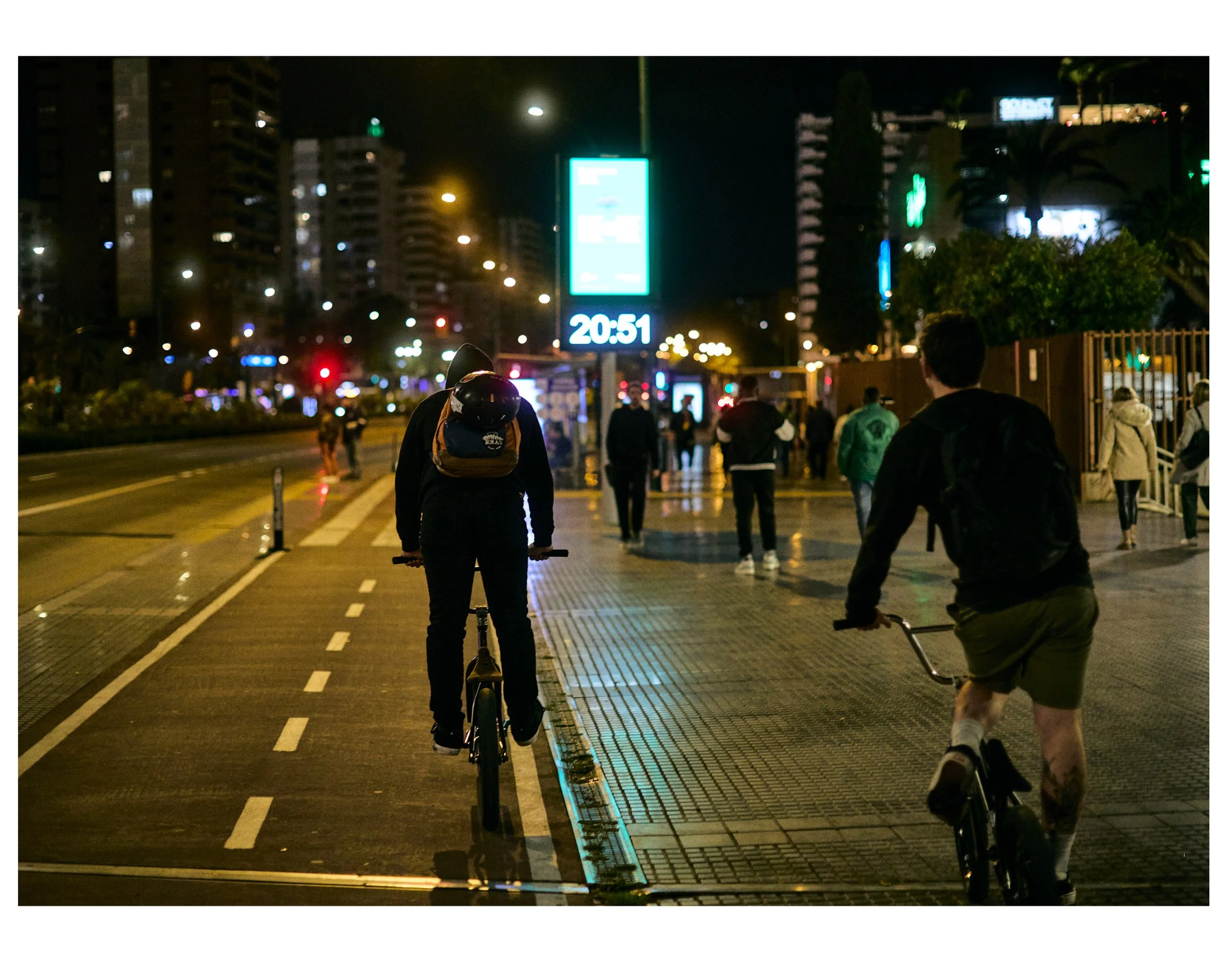 People riding bikes and walking on a city sidewalk at night with illuminated buildings and a digital clock showing 20:51.