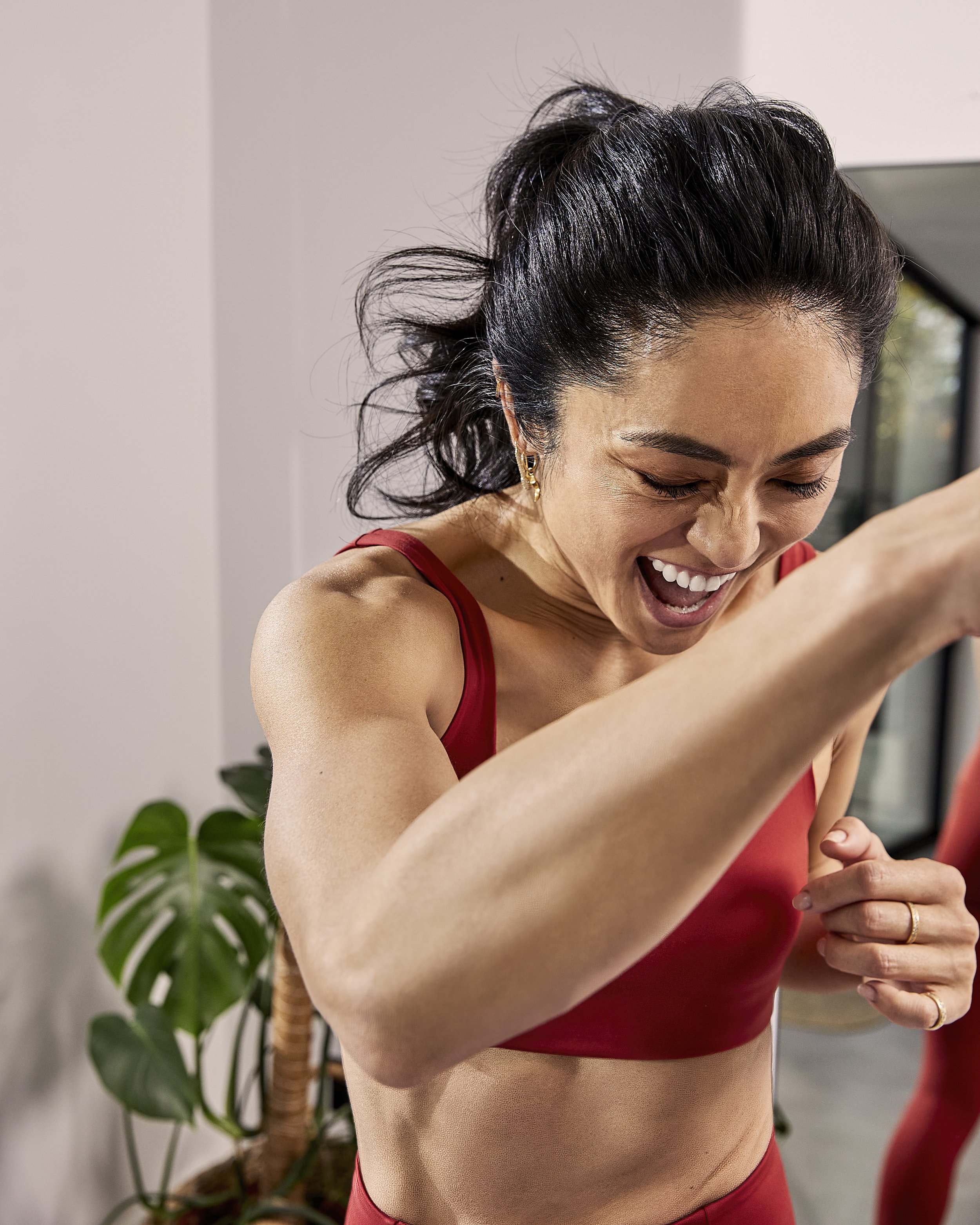 A woman wearing a red sports bra and shorts is smiling and laughing while looking down, with her right arm raised and her left hand grasping her right wrist, in a bright indoor setting with a plant in the background.