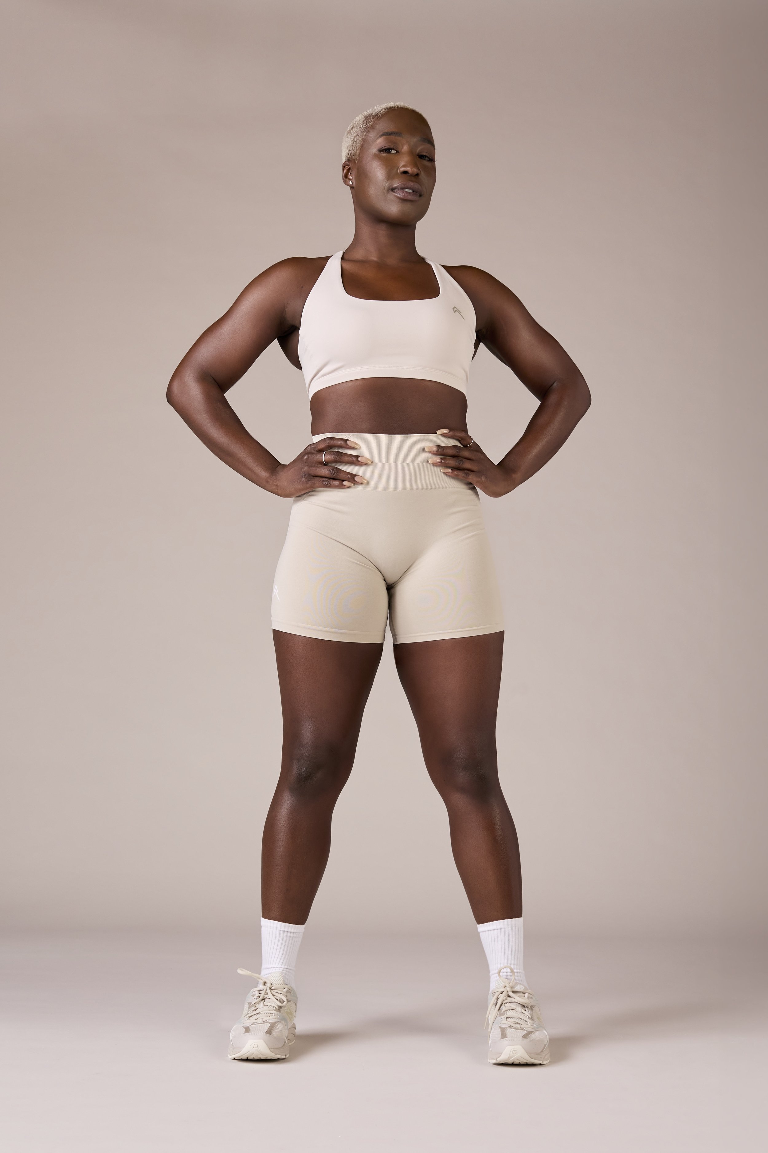 A confident woman with short blond hair wearing a white sports bra, beige shorts, and sneakers standing with hands on her hips.