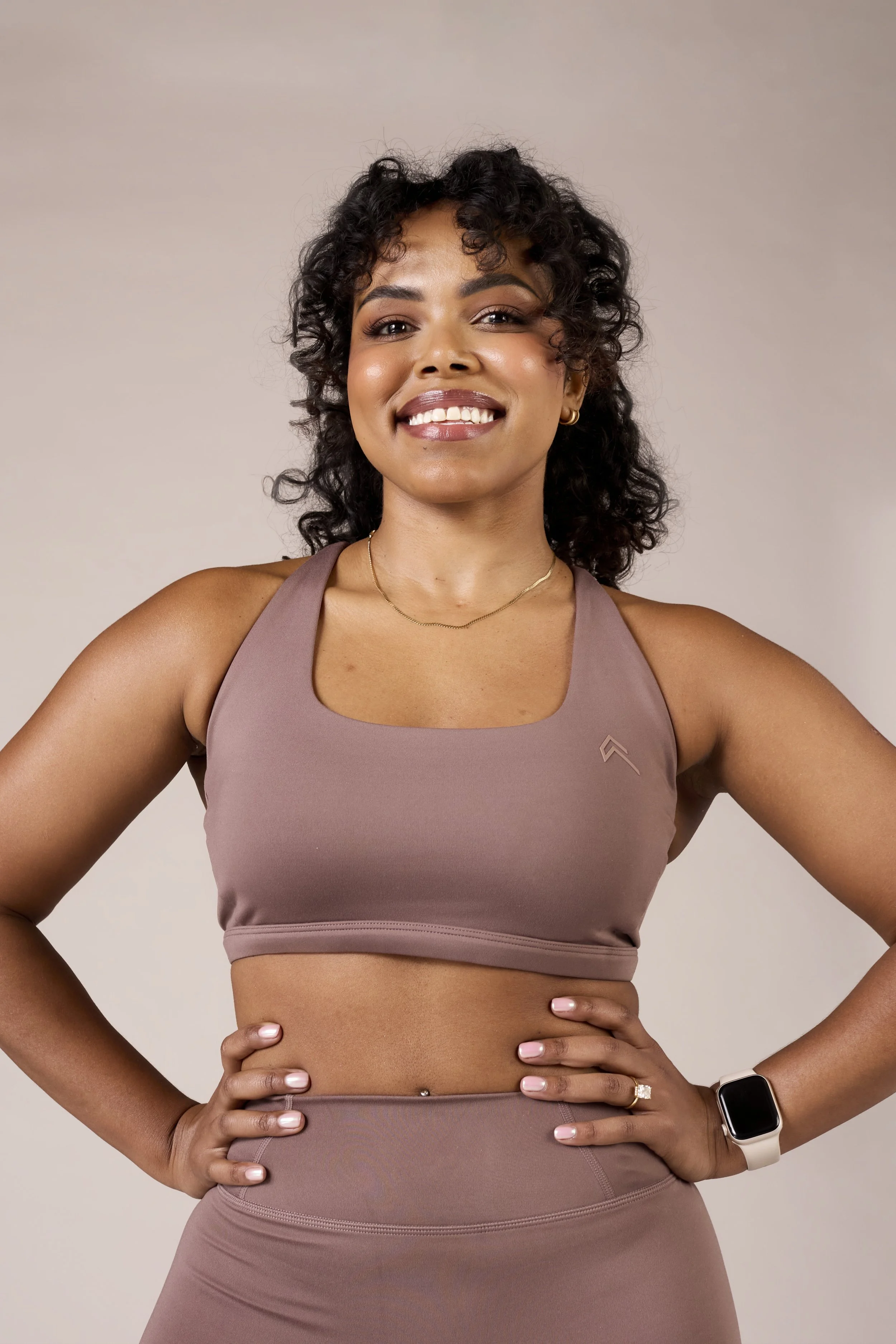 Woman in athletic wear smiling with hands on hips, wearing a smartwatch and jewelry, posing against a neutral background.