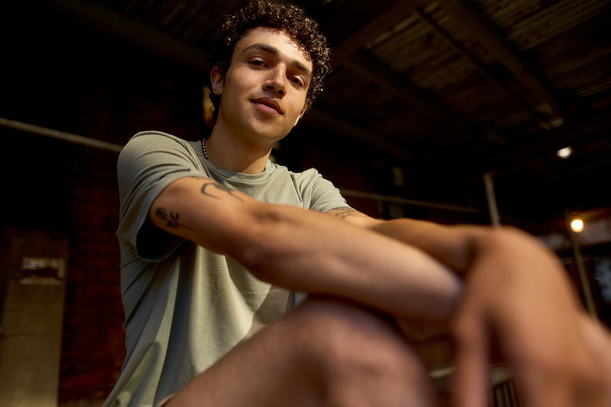 Young man with curly hair and tattoos on his arm, wearing a light gray T-shirt, leaning forward with a confident expression and a slight smile inside a dimly lit industrial-style space.