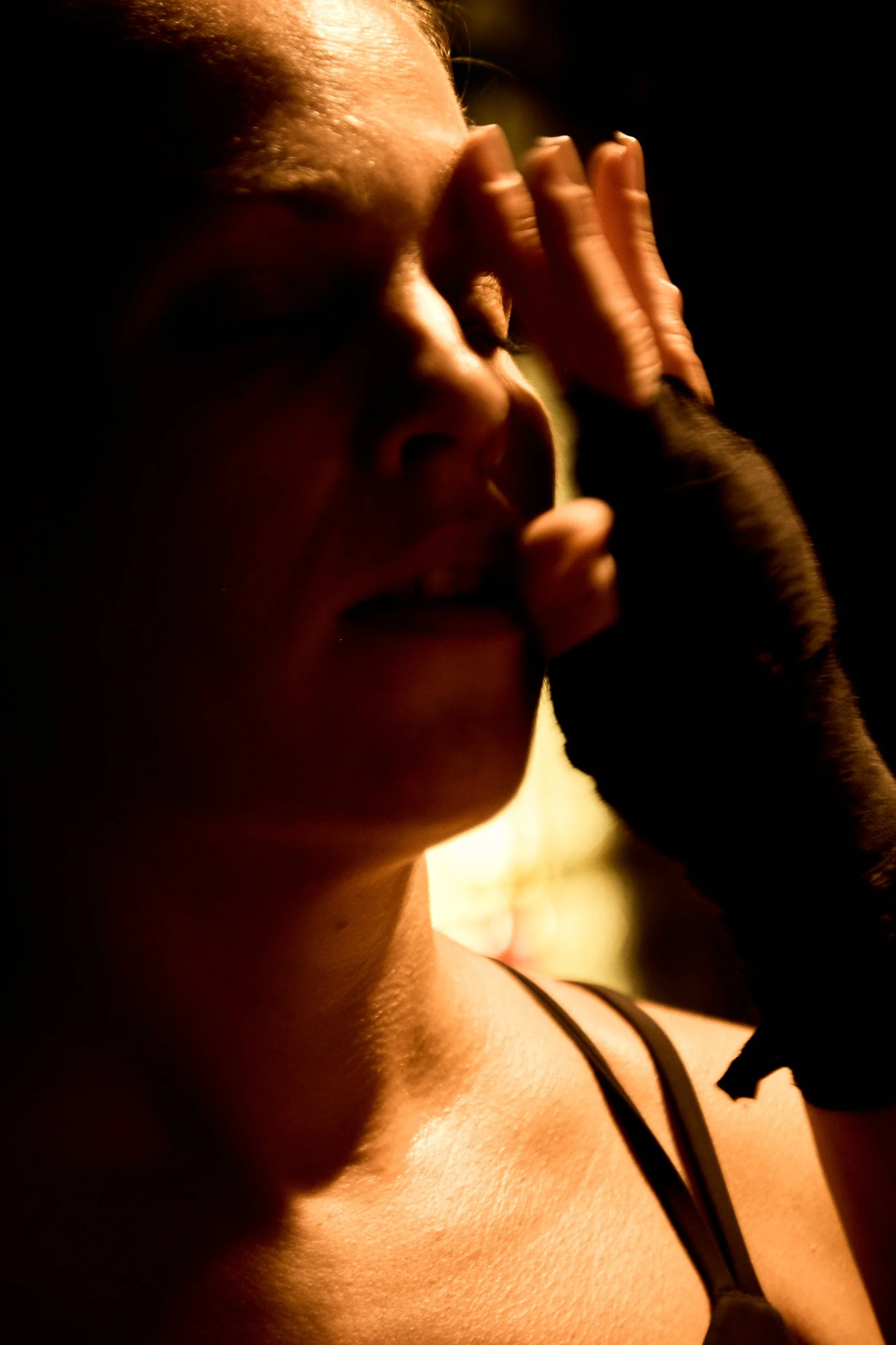 Close-up of a woman with short hair, eyes closed, face etched with emotion, resting her hand on her forehead with a black glove, in dramatic lighting.