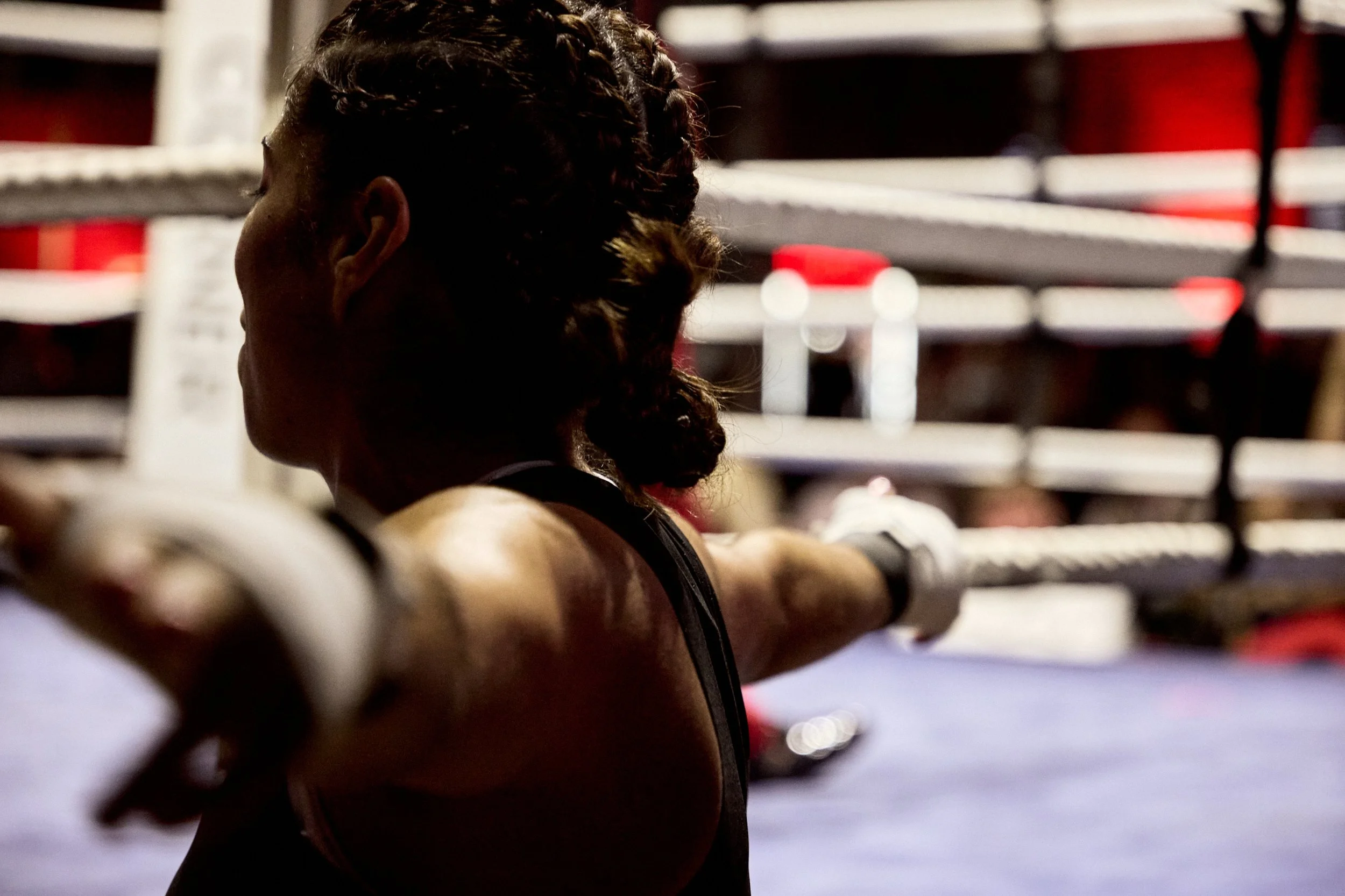 Female boxer resting against ropes inside a boxing ring, facing away from the camera.