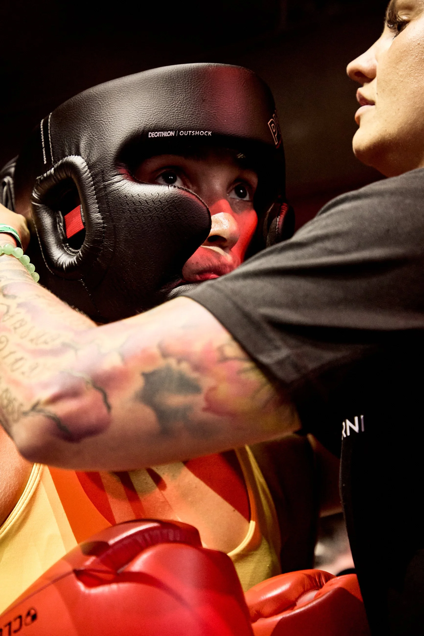 A female boxer wearing a black headgear, looking focused during a match with her coach or trainer adjusting her headgear. The trainer's tattooed arm is visible.