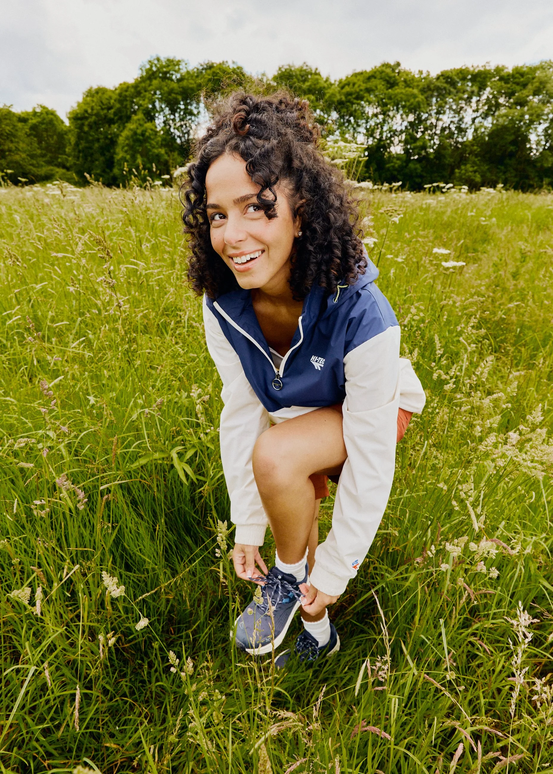 A young woman with curly dark hair is outdoors in a grassy field, smiling and tying her shoelaces. She is wearing a blue and white windbreaker, shorts, and sneakers.