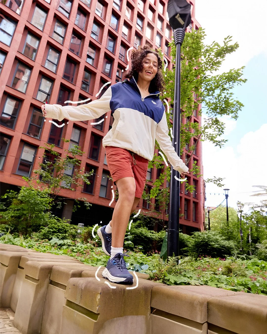 A young woman balancing on a low stone wall in an urban park, wearing a color-block windbreaker, red shorts, and athletic shoes, with a tall red-brick building in the background.