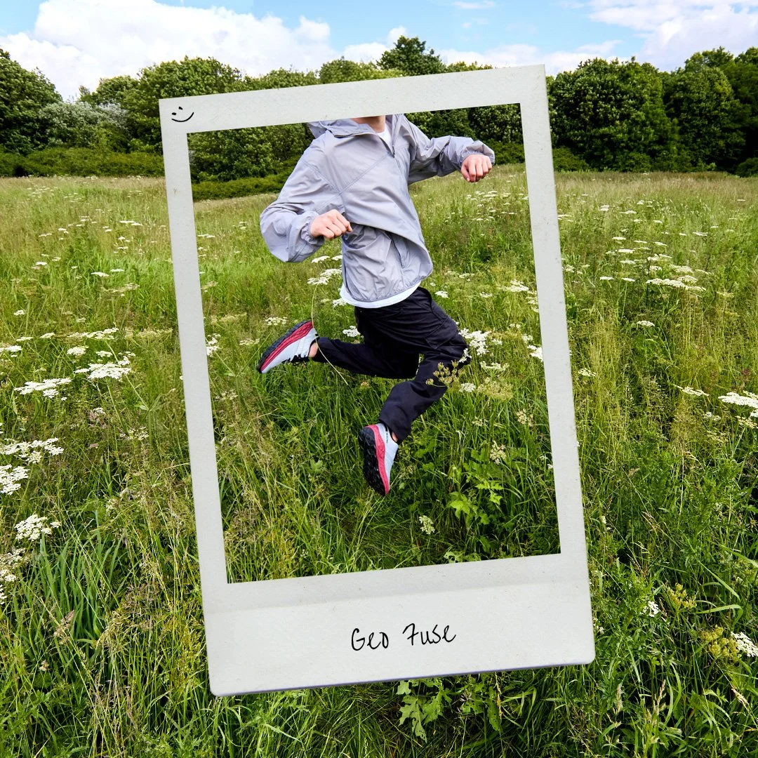 Person jumping in a grassy field with wildflowers, seen through a large white frame with a smiley face drawn on the top left corner, and a handwritten label 'Geo Fuse' at the bottom.