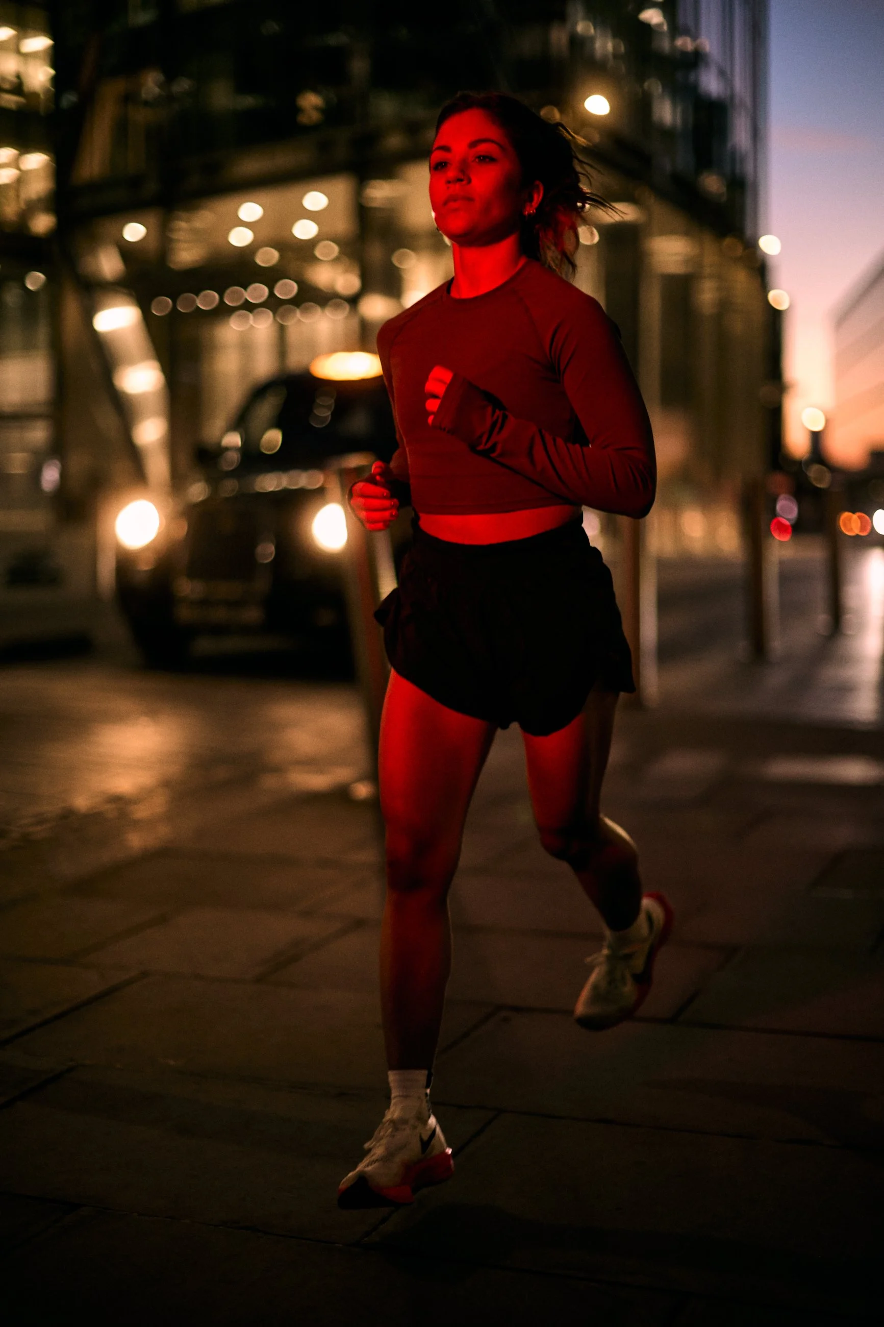 A woman running outdoors at dusk or night, illuminated by streetlights, wearing a long-sleeve athletic top, shorts, and running shoes.