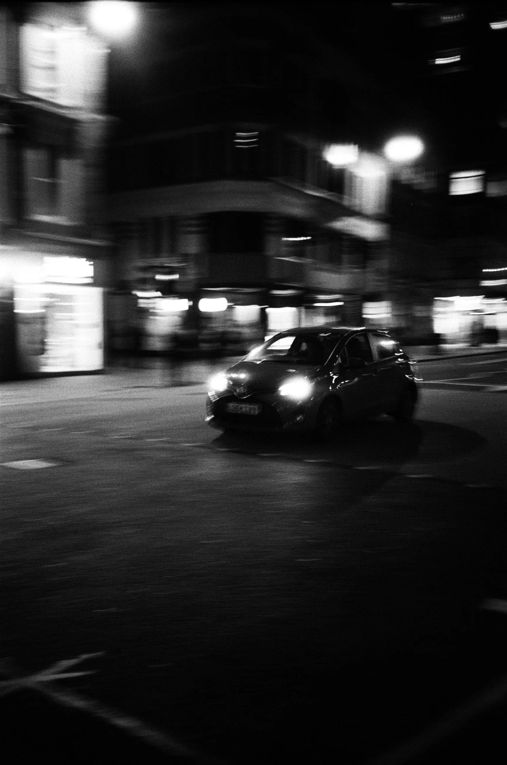 A black and white photo of a car driving on a city street at night, with blurred lights and buildings in the background.