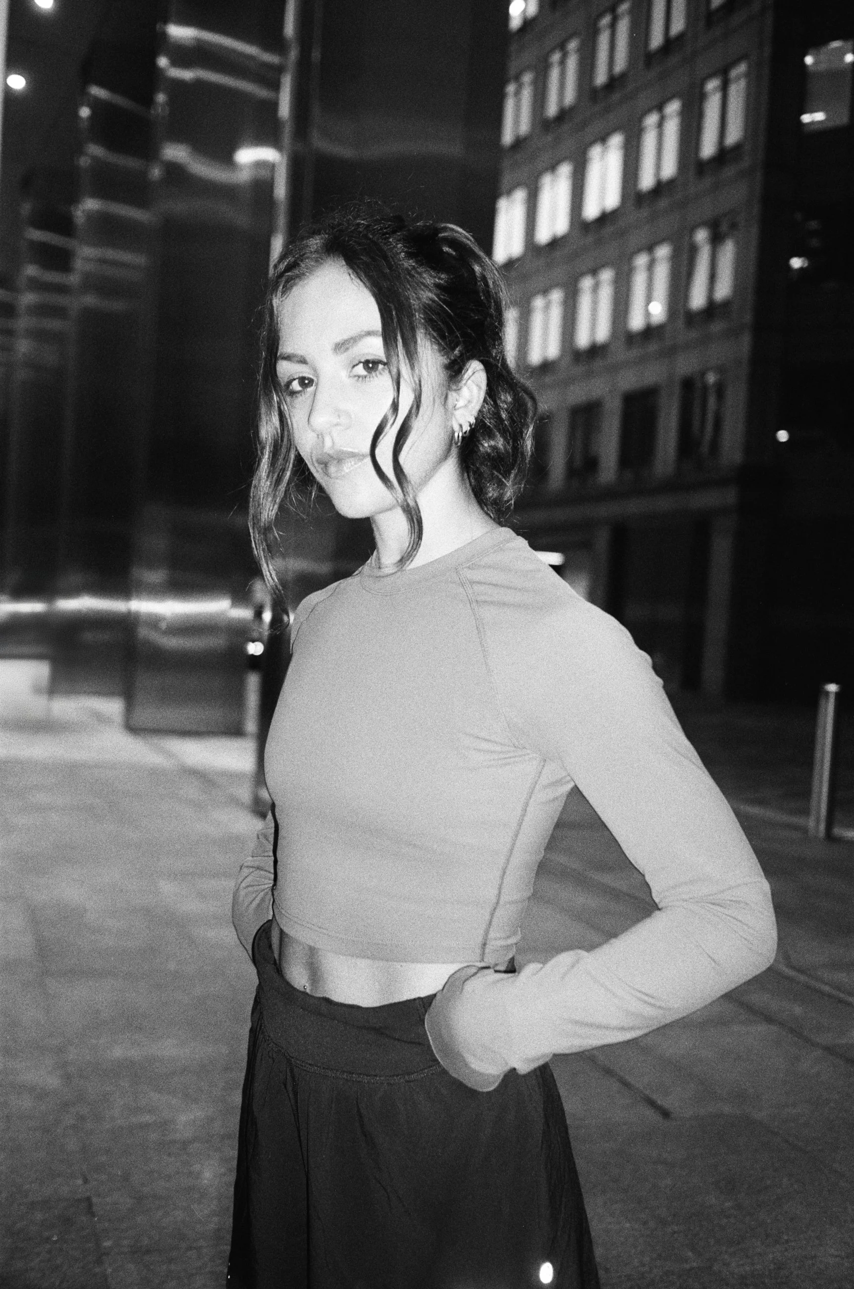 A young woman with dark hair styled in loose waves, wearing a crop top and a skirt, standing on a city sidewalk at night in black and white.