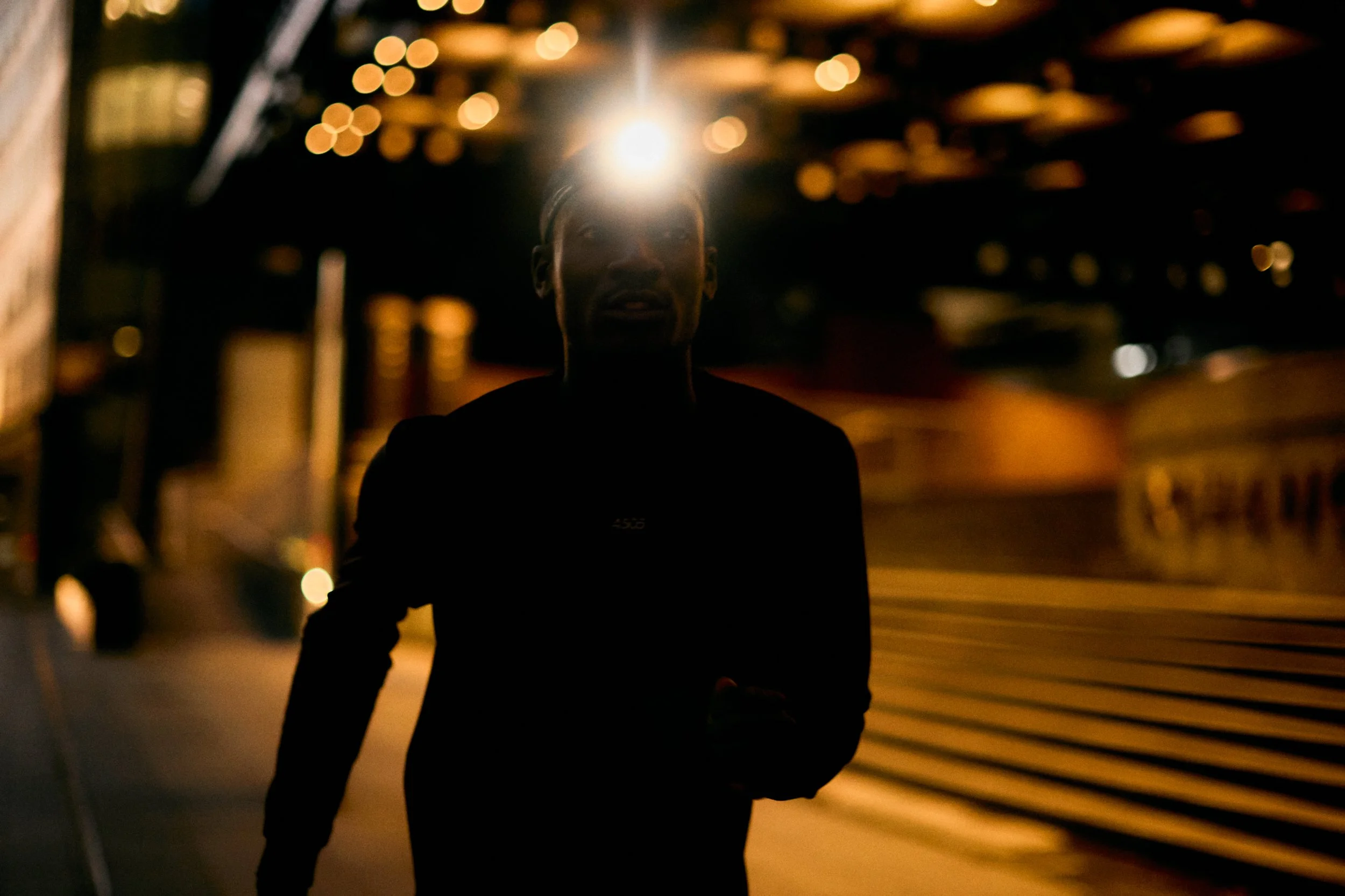 A person running in a dark urban environment at night, with a bright light shining behind their head creating a silhouette effect.
