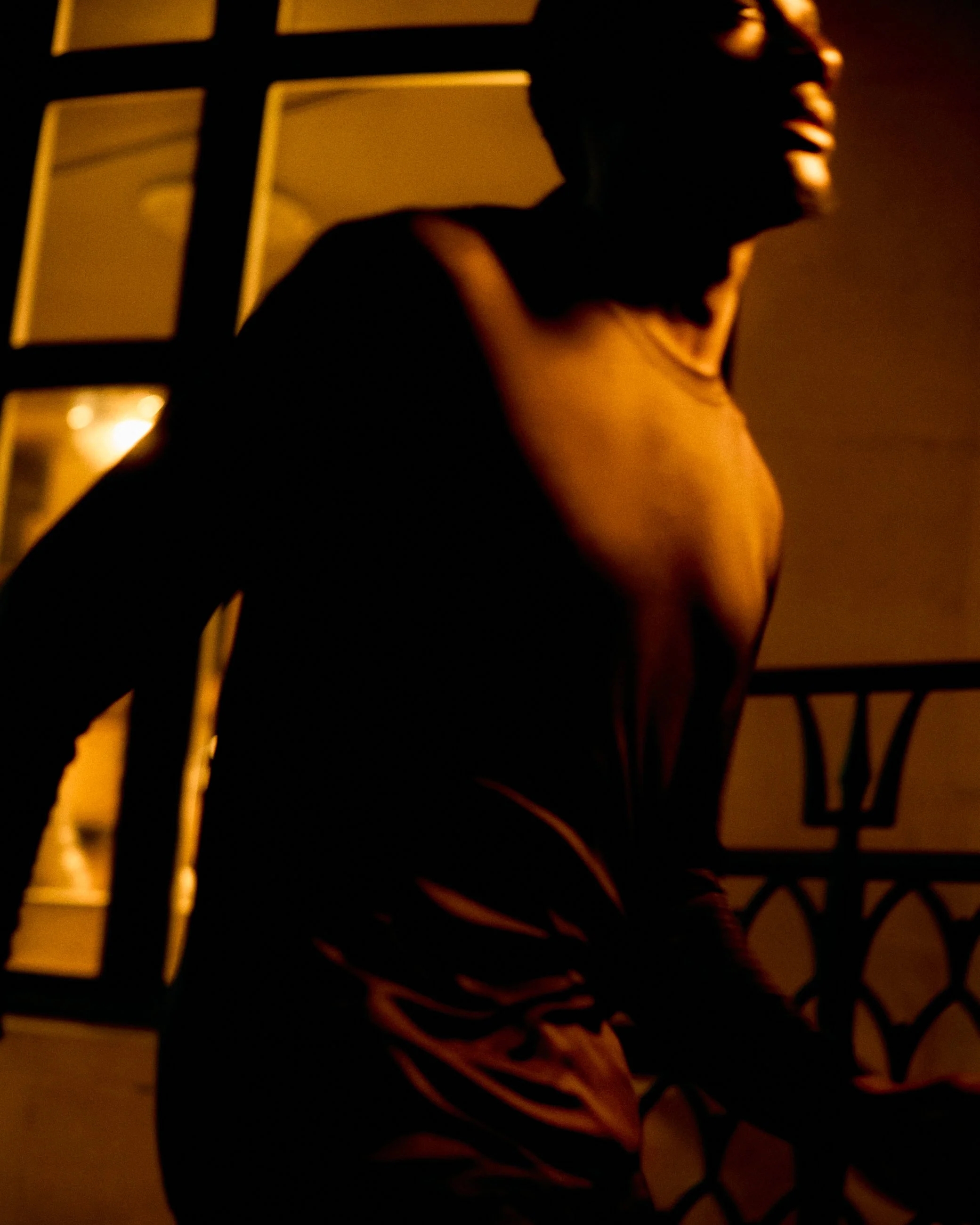 A person with short hair sitting on a balcony at night, illuminated by warm lighting, with a large window and decorative metal railing in the background.
