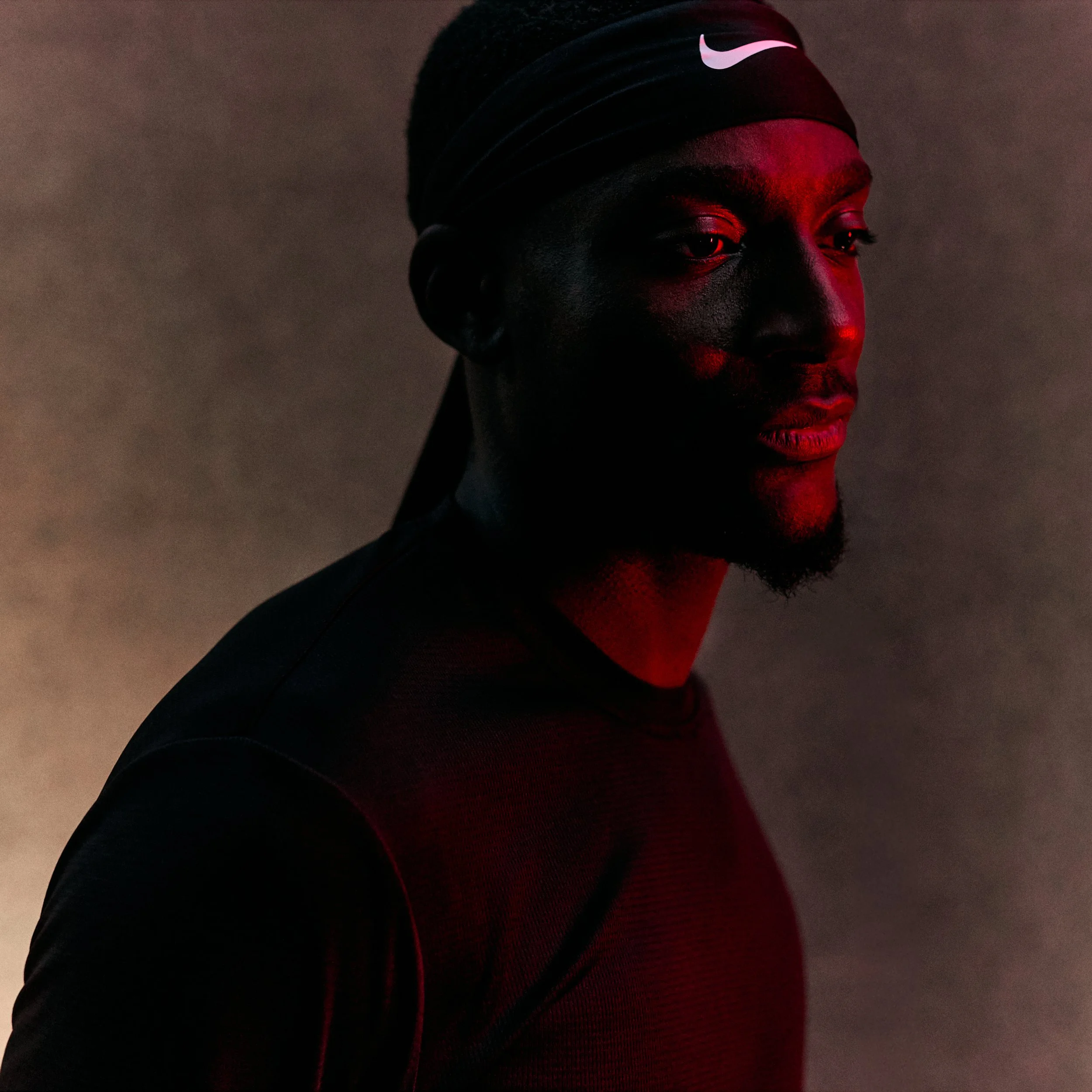 Portrait of a man wearing a black Nike headband, with dark skin and facial hair, looking to the side against a dark background with red lighting.